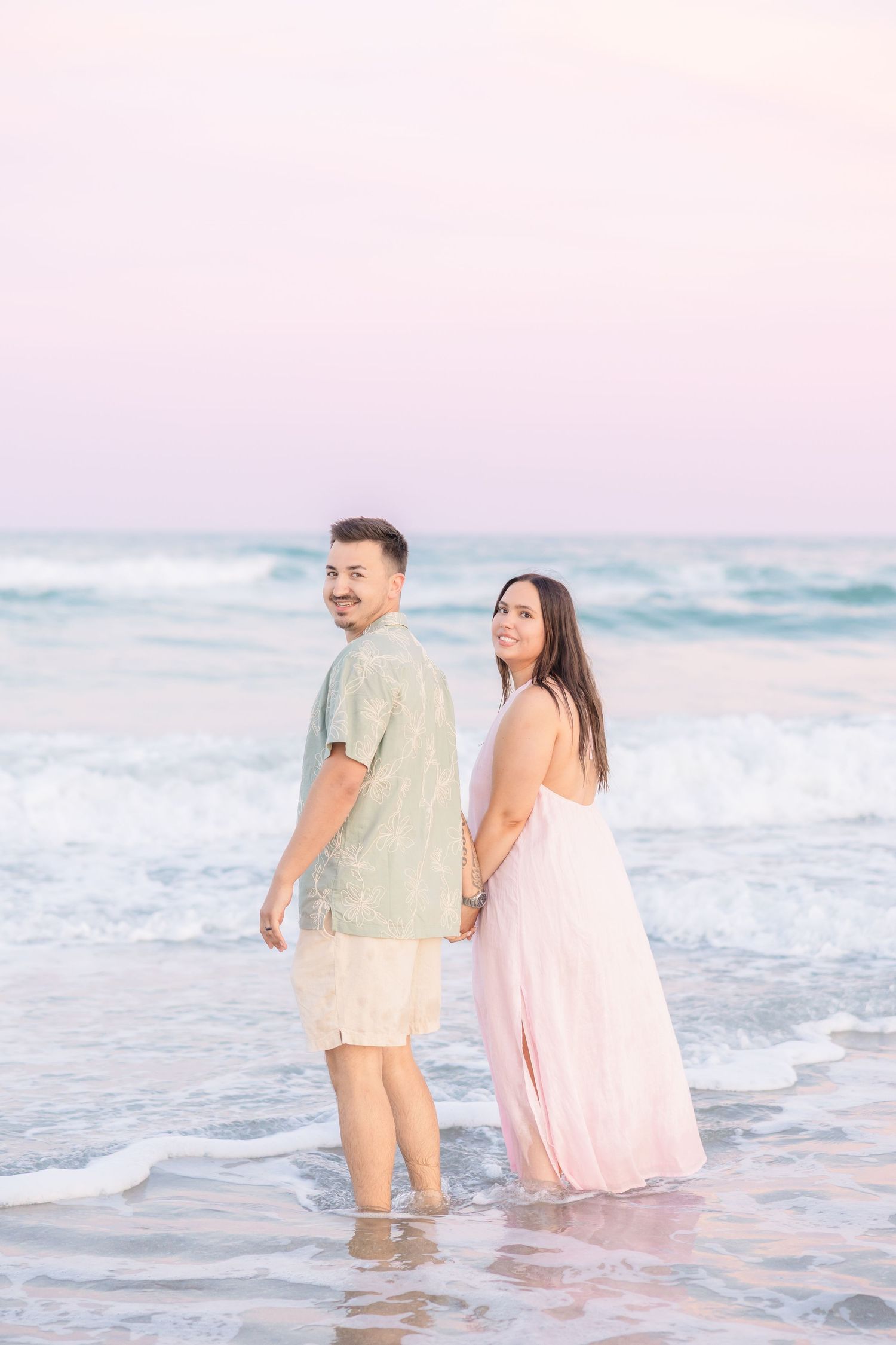 Couple holding hands at beach during sunset, woman in pink dress and man in green shirt standing in ocean waves.