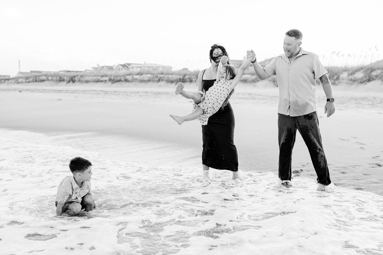 Black and white beach photo of parents playfully swinging their daughter while son plays in sand.