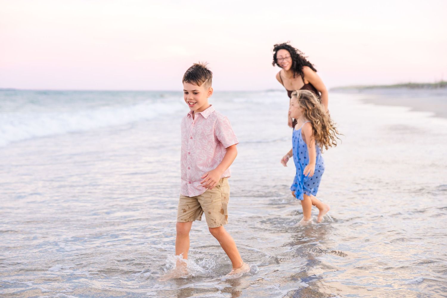 A mother and two children laugh and play in shallow ocean waves at sunset.