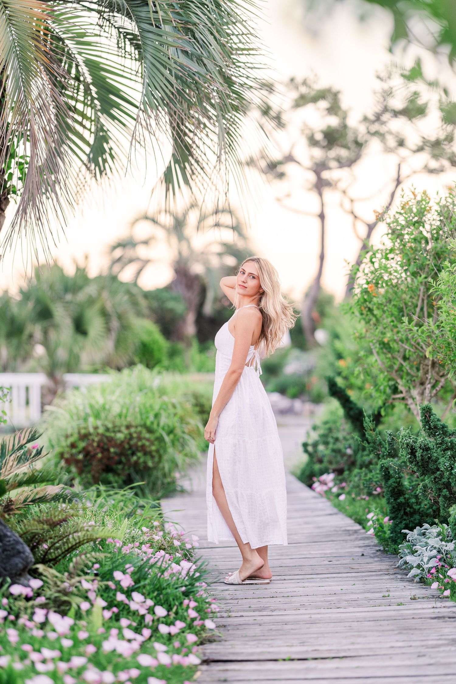 A person in a flowing white dress twirls gracefully on a wooden boardwalk surrounded by tropical foliage and pink flowers.