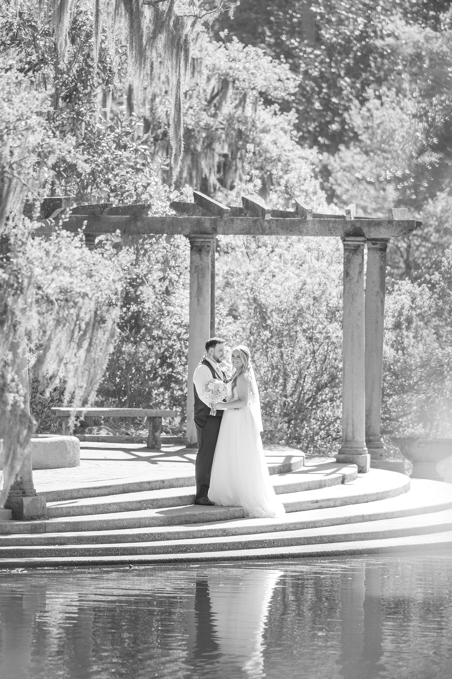 Bride and groom share a romantic kiss under a stone pergola by a reflective pond in black and white in Airlie Gardens in Wilmington, NC
