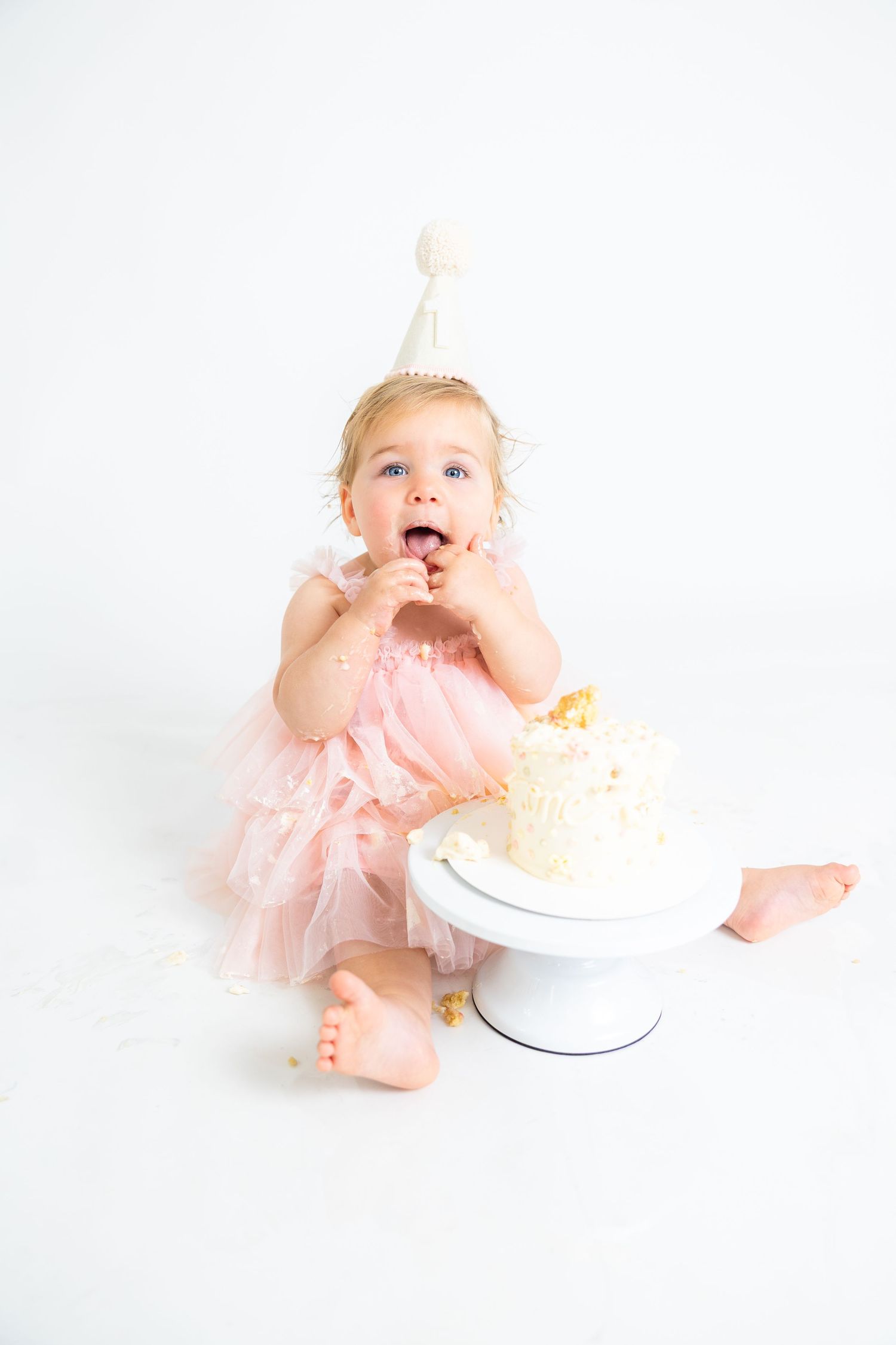 Baby in pink dress and party hat tasting frosting during cake smash photo session.