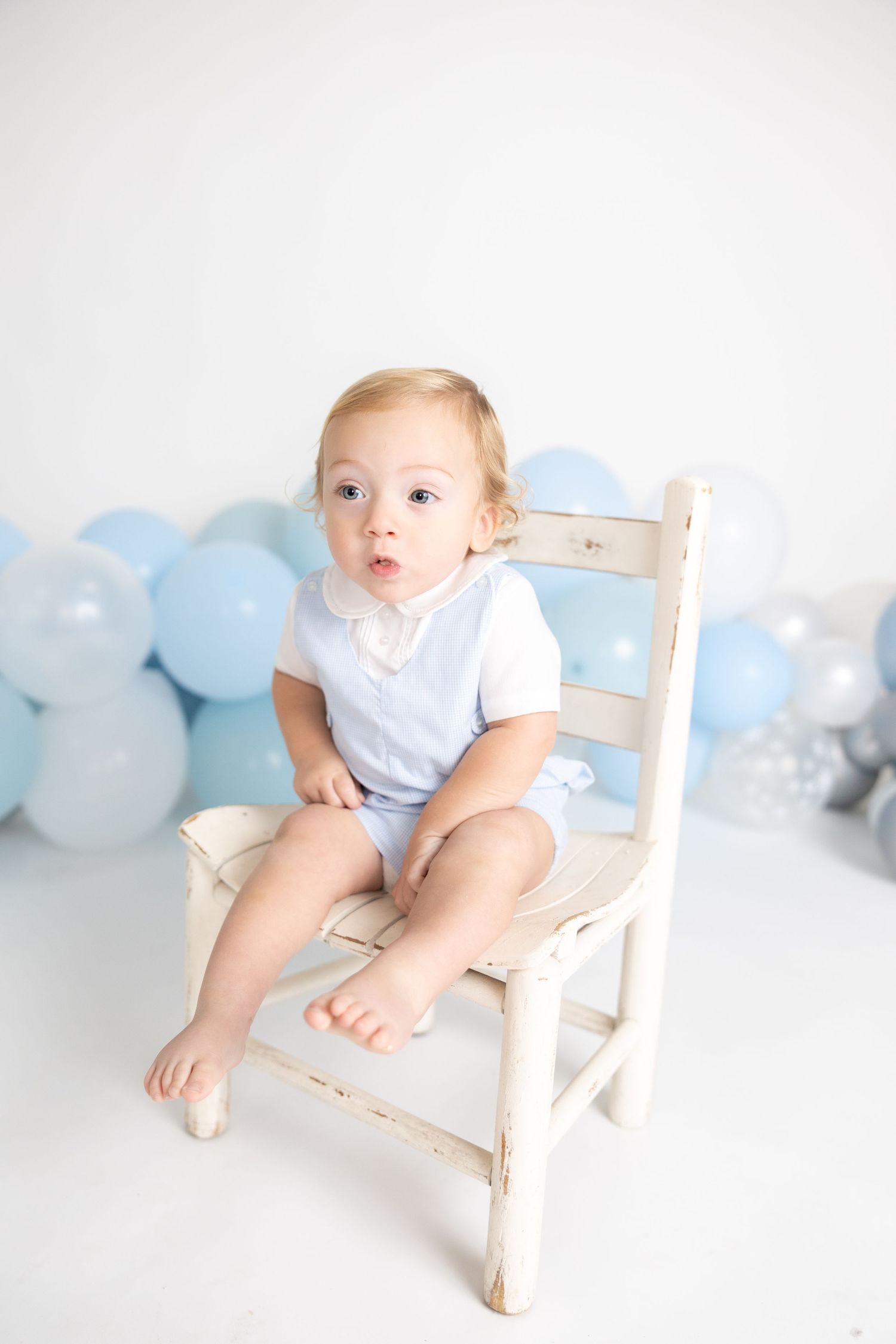 Baby sitting on white wooden chair with blue balloon decoration behind.