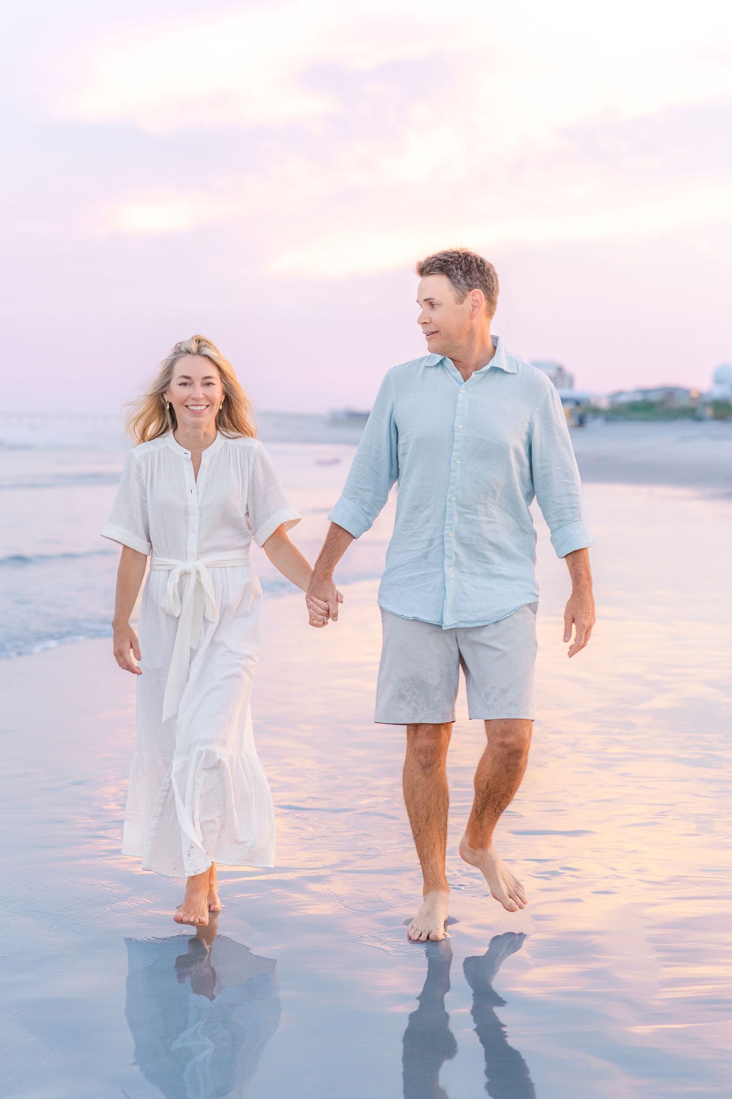 A couple walking hand in hand on a beach at sunset wearing coordinating white and blue outfits.