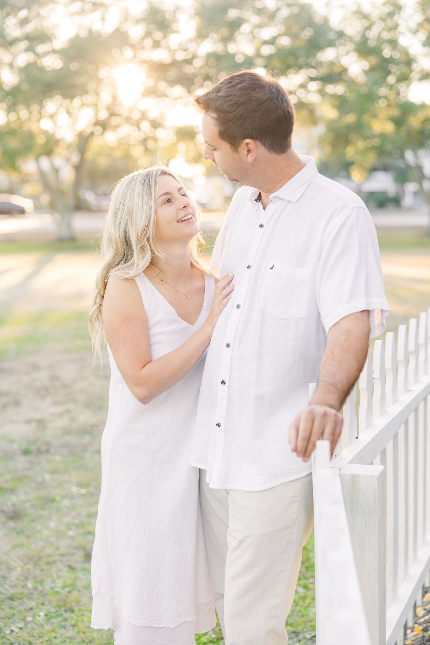 Two people share intimate moments while leaning against a white fence during a golden sunset.