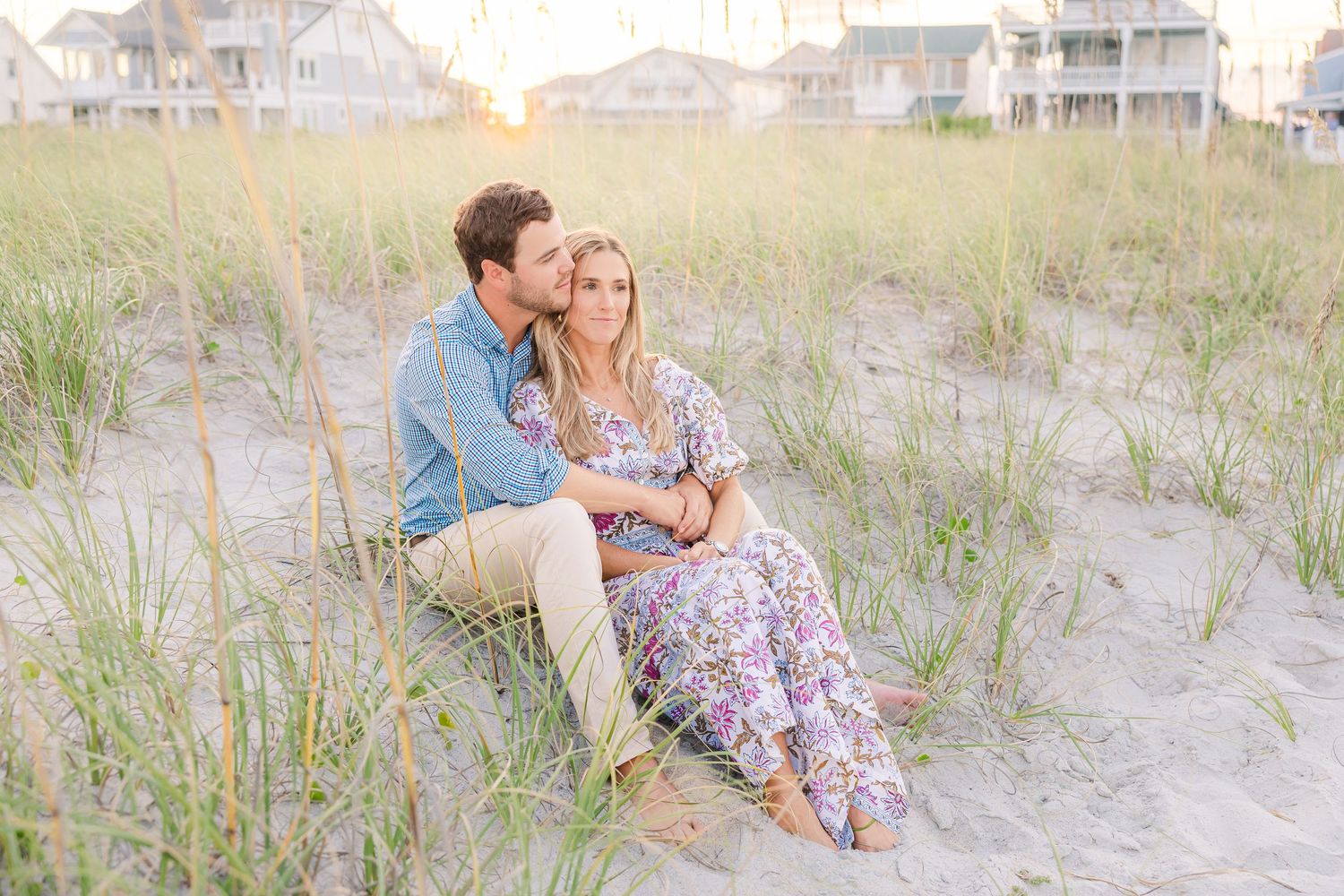 Couple relaxing on beach dunes during golden hour with coastal homes behind.