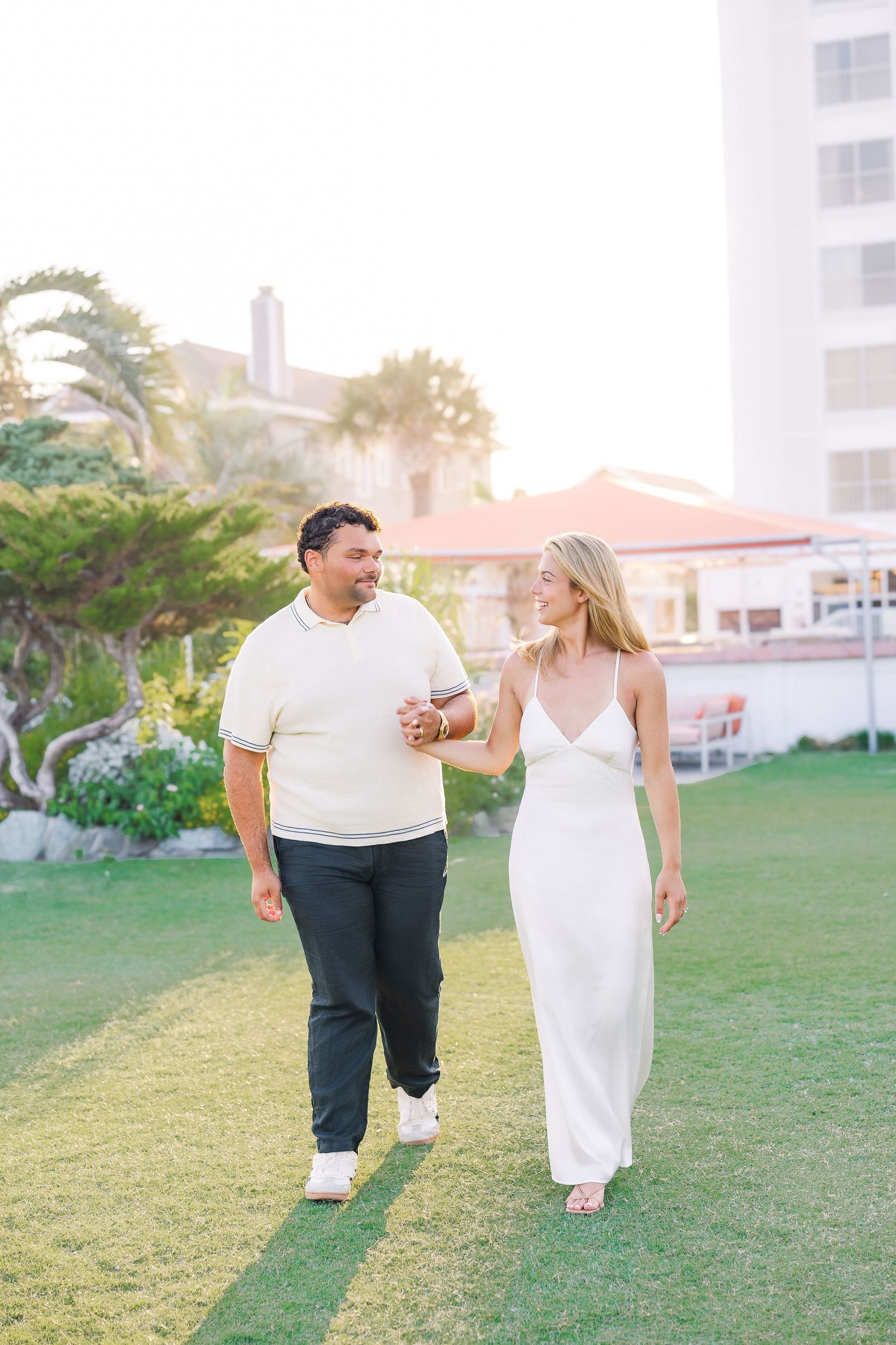 Couple walks hand-in-hand on green lawn at golden hour near a resort with palm trees.