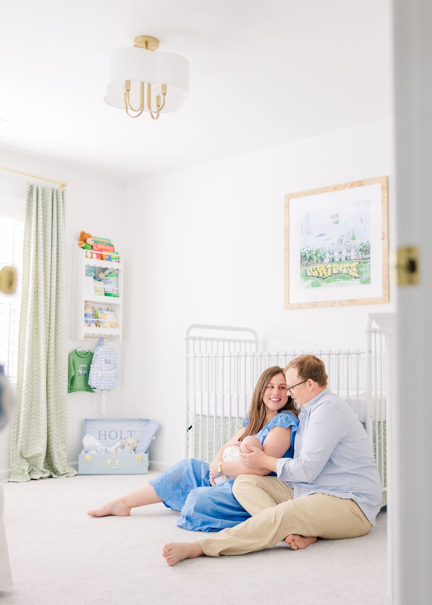 Parents sit on nursery floor with newborn, white crib and watercolor art behind them in a bright, elegant room.