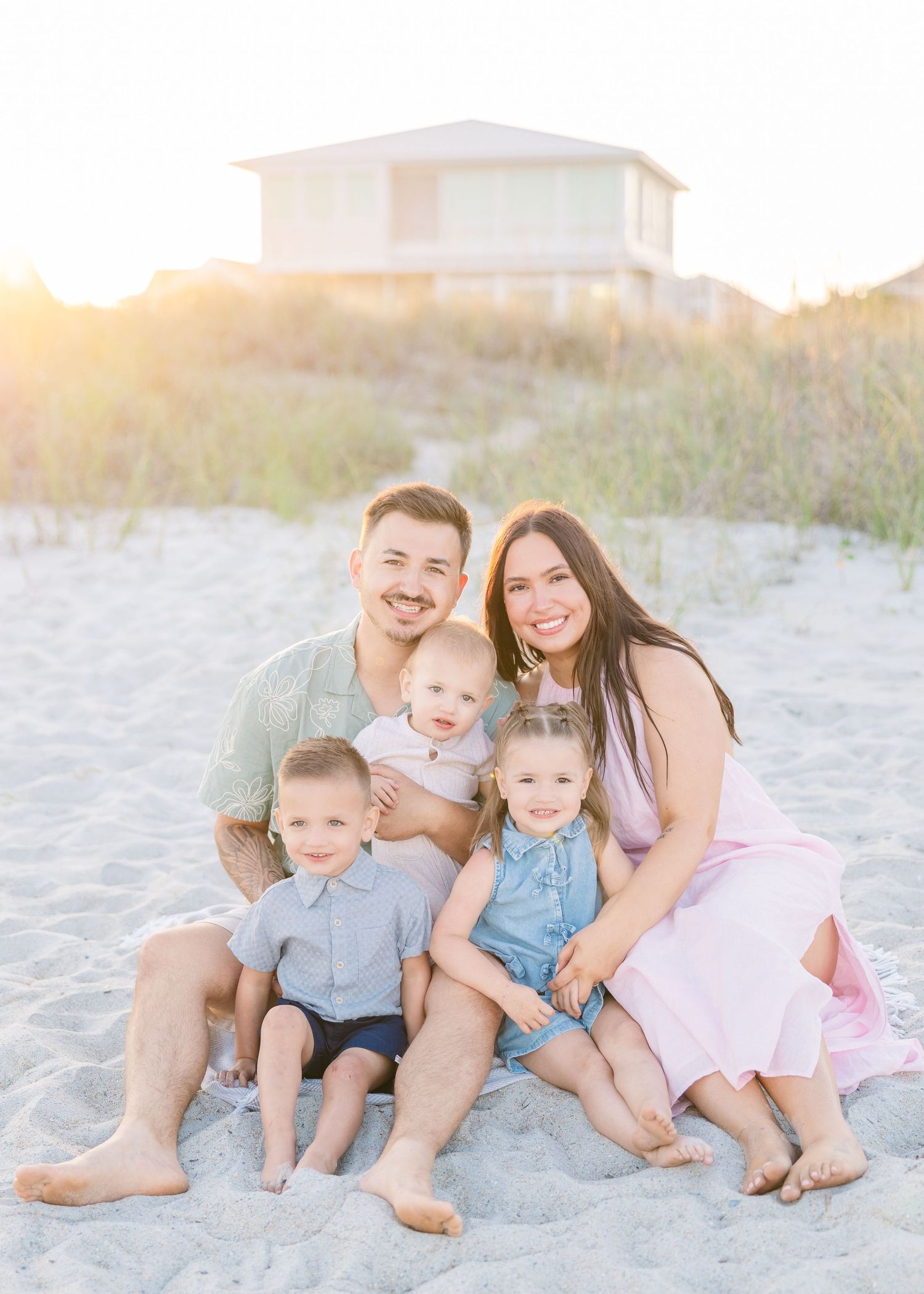 Happy family of five poses on sandy beach at golden hour with coastal house in background.