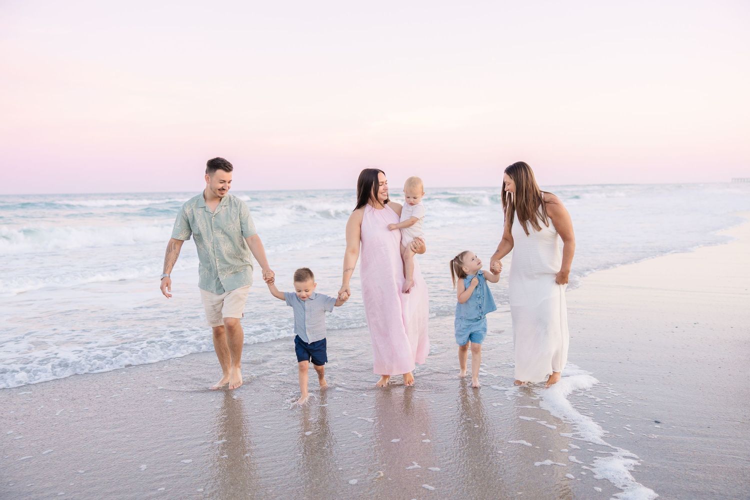 Happy family of six walking along a pastel sunset beach, holding hands near the ocean waves.