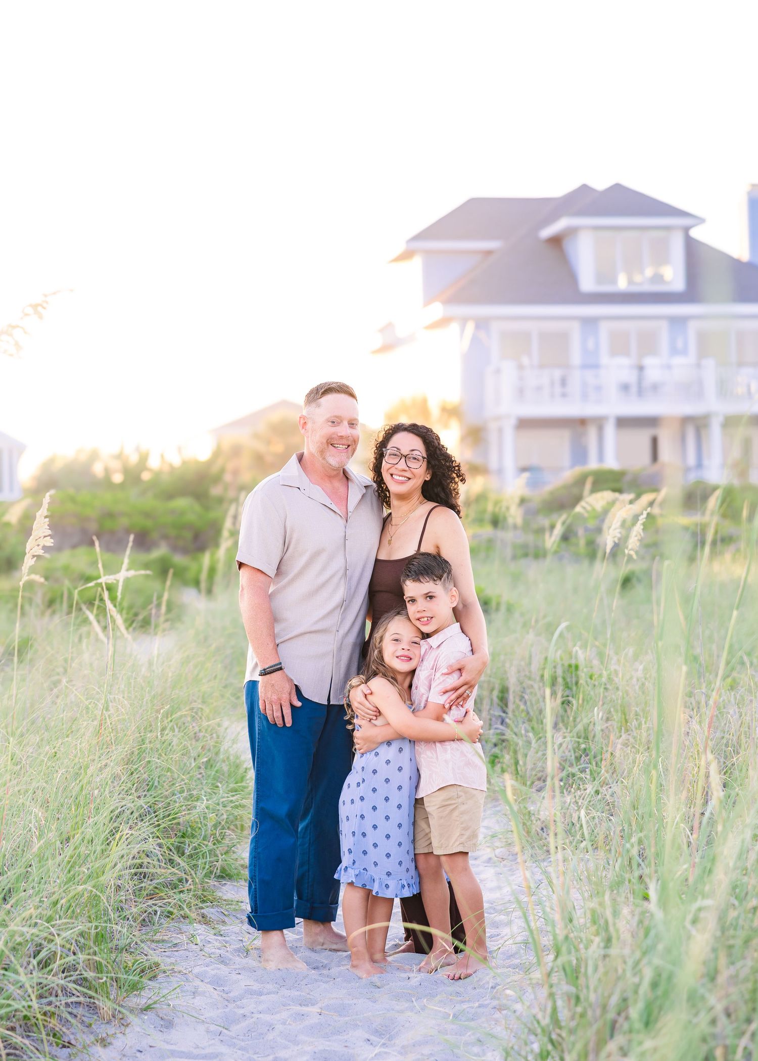 Family of four poses on sandy beach path at golden hour with sea oats and coastal home backdrop.