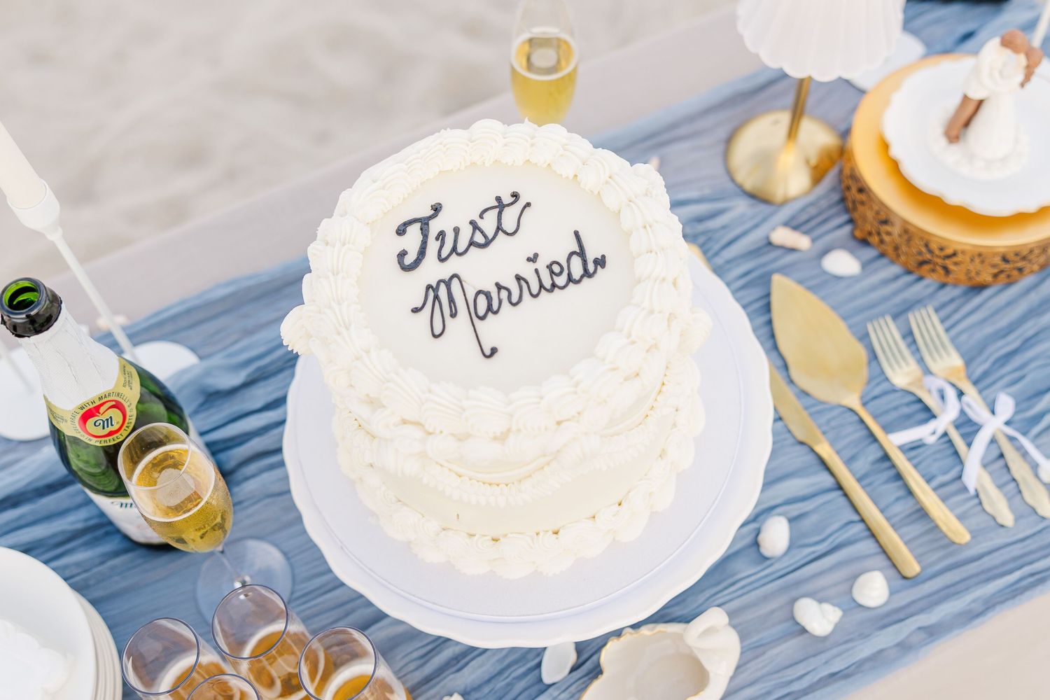 White wedding cake reading Just Married displayed on blue table with gold utensils at beach reception in Carolina Beach near Wilmington, NC.