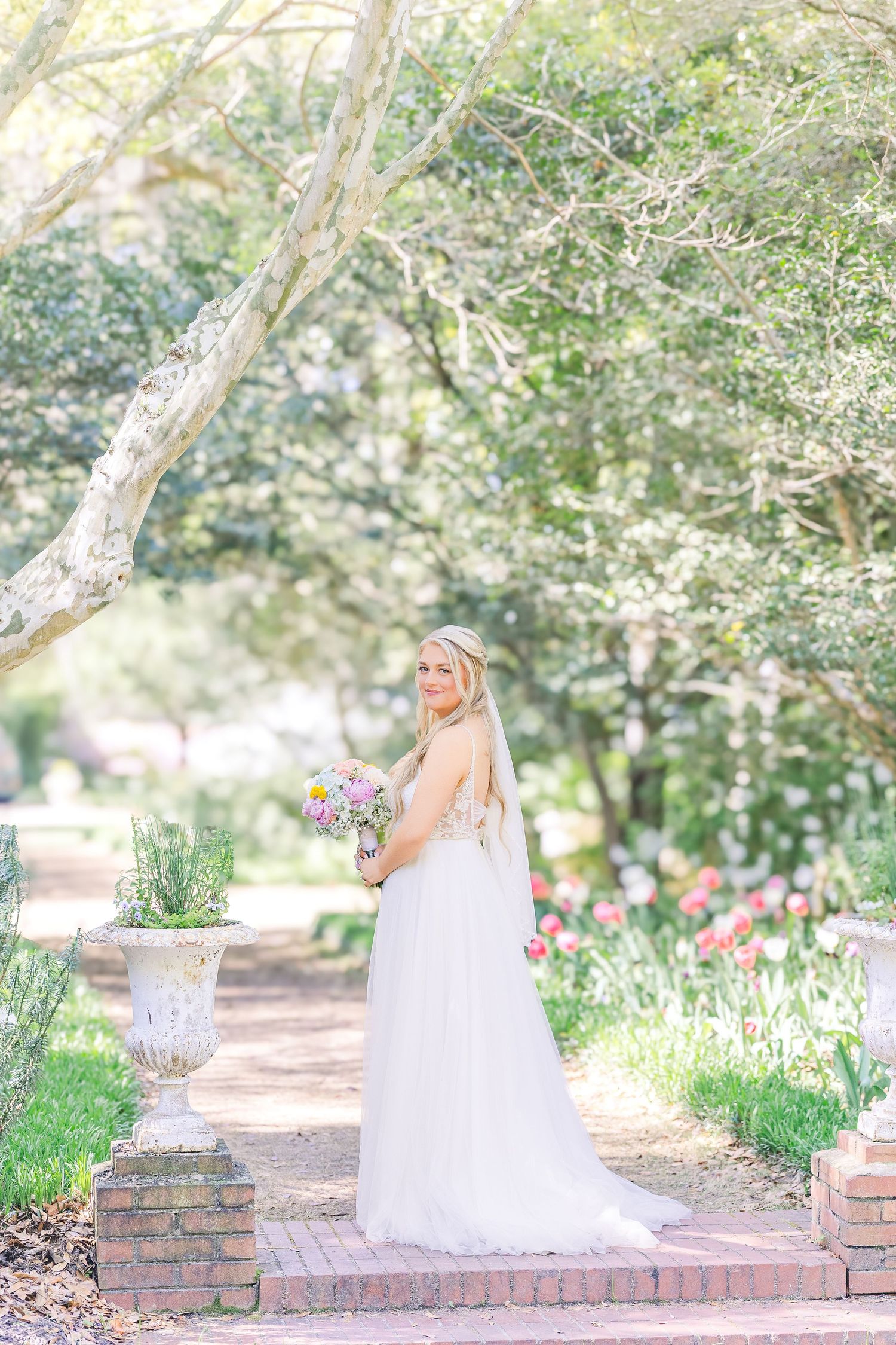 Bride in white gown holding colorful bouquet stands on brick path in lush garden with tulips and trees in Airlie Gardens in Wilmington, NC