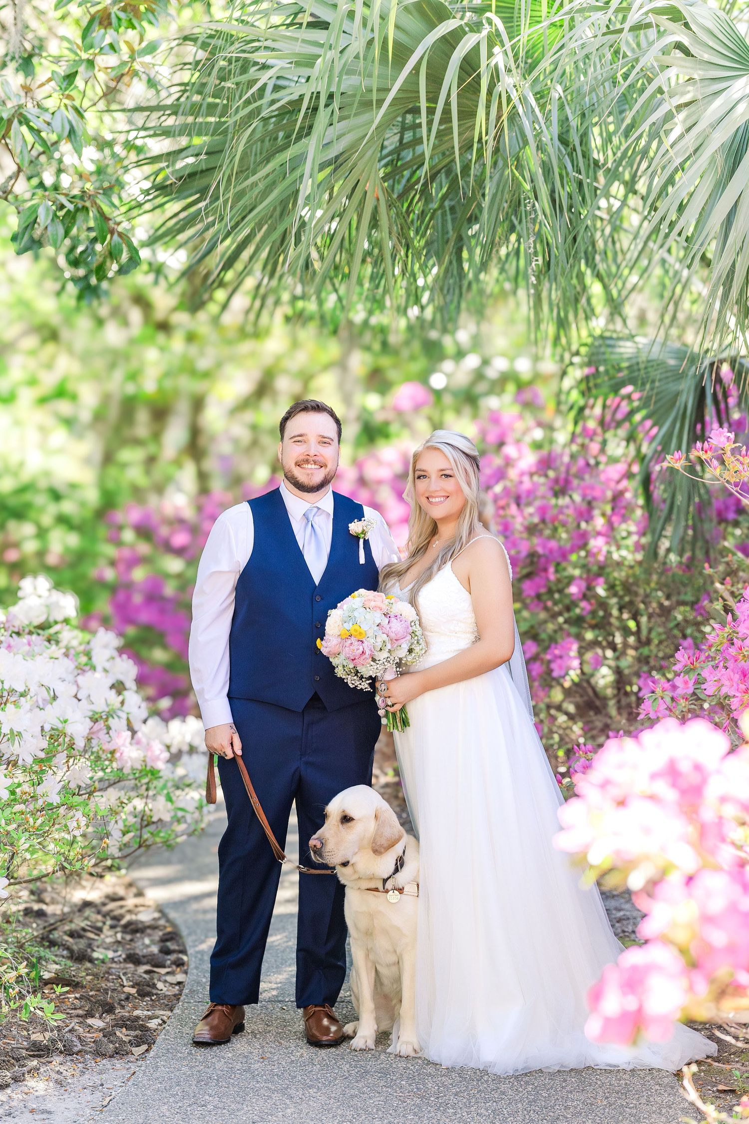 Bride and groom pose with golden retriever dog amid colorful blooming flowers and palm trees on wedding day in Airlie Gardens in Wilmington,