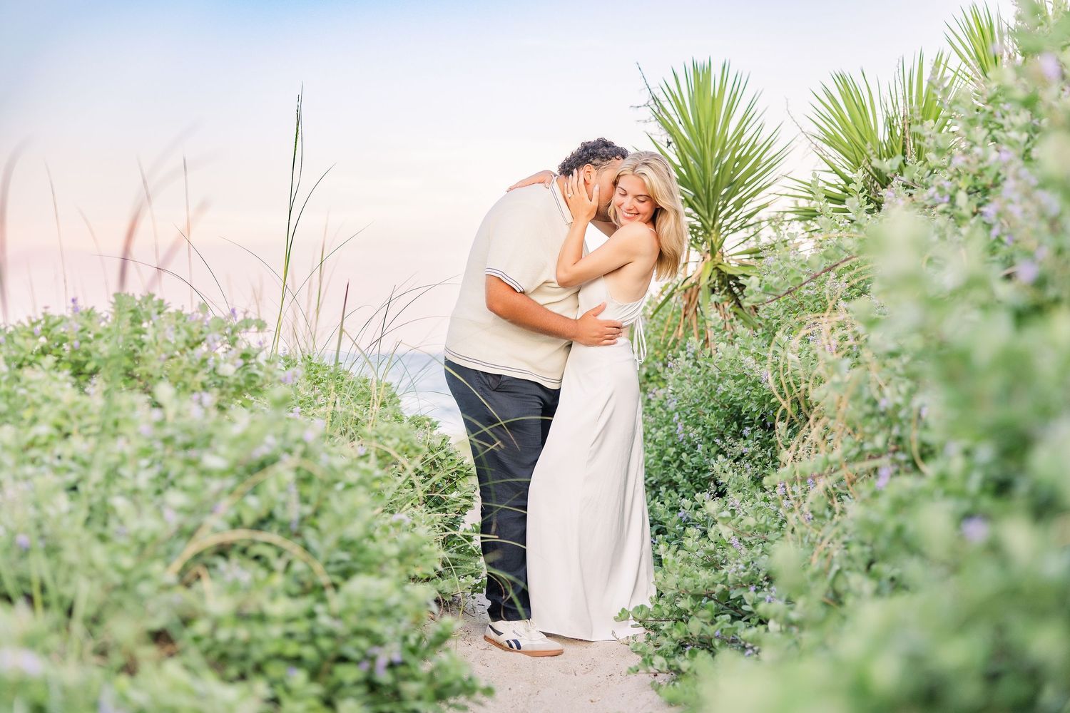 Couple embracing and kissing surrounded by lush coastal greenery and sea oats at golden hour.