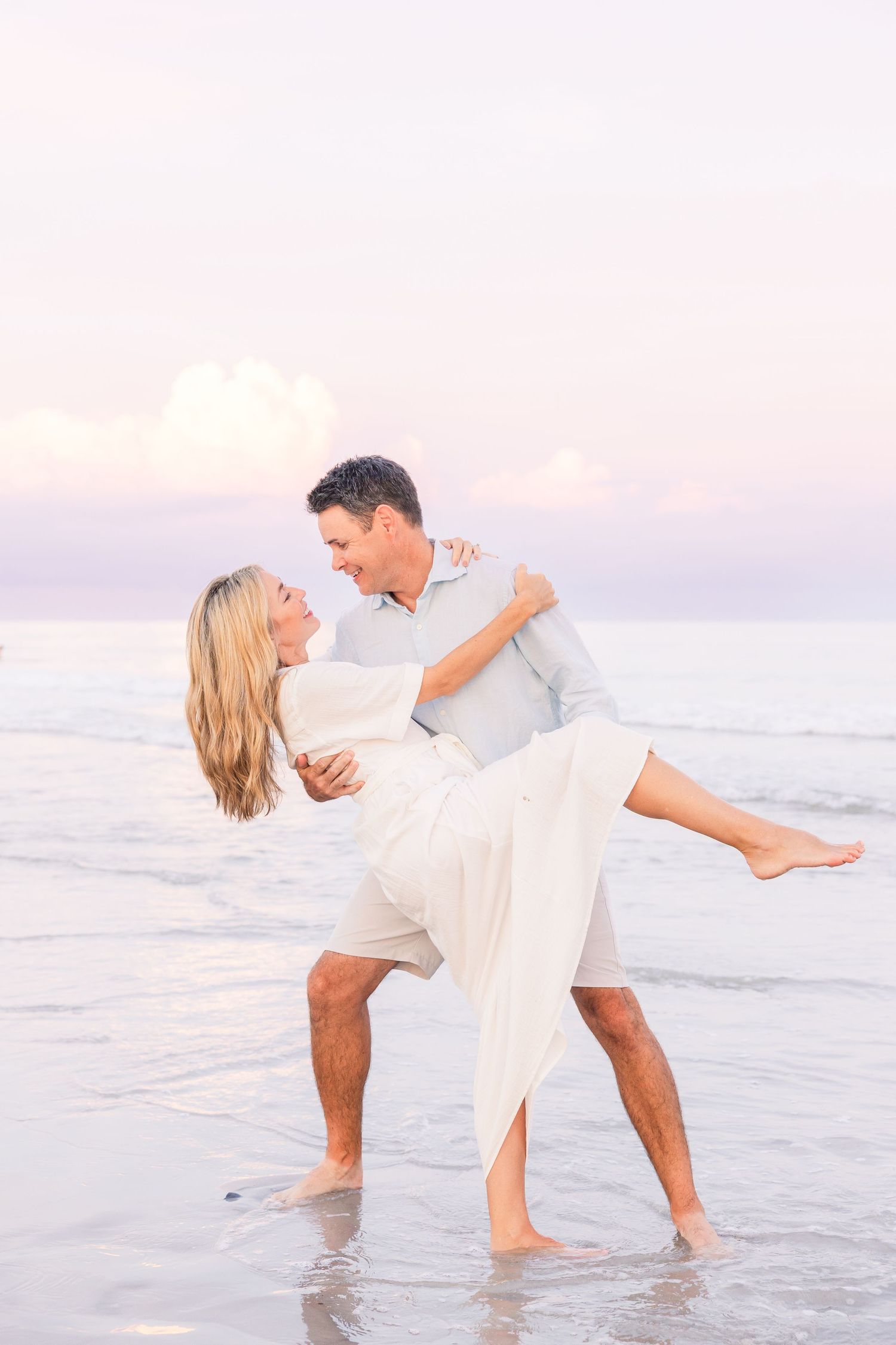 A romantic couple dances and embraces in white clothing at the beach during sunset.