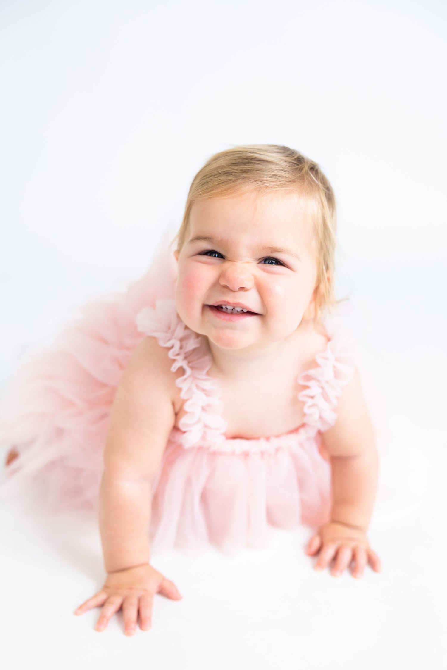 Happy baby wearing pink tulle dress crawling on white backdrop.