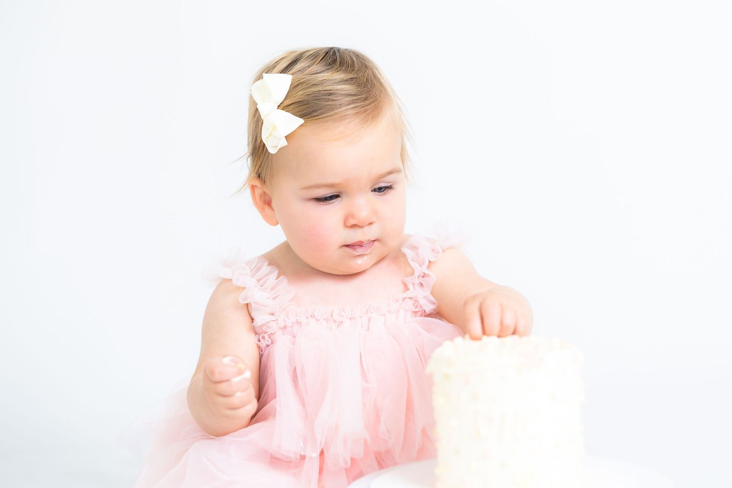 Baby wearing pink dress reaches toward white cake stand in studio setting.