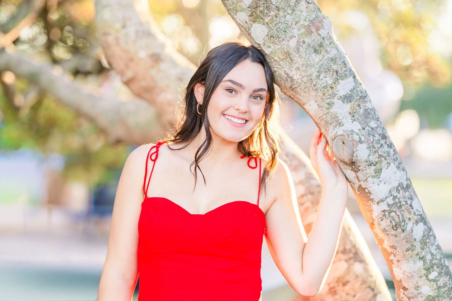 Outdoor portrait in red dress leaning against tree with sunlit background.