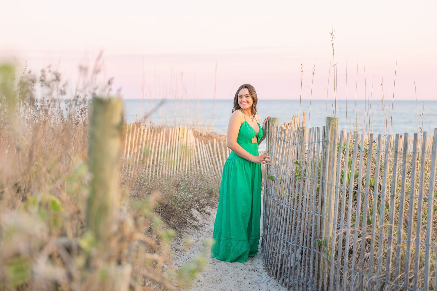 Beach portrait featuring a flowing emerald dress against a weathered wooden fence at sunset.