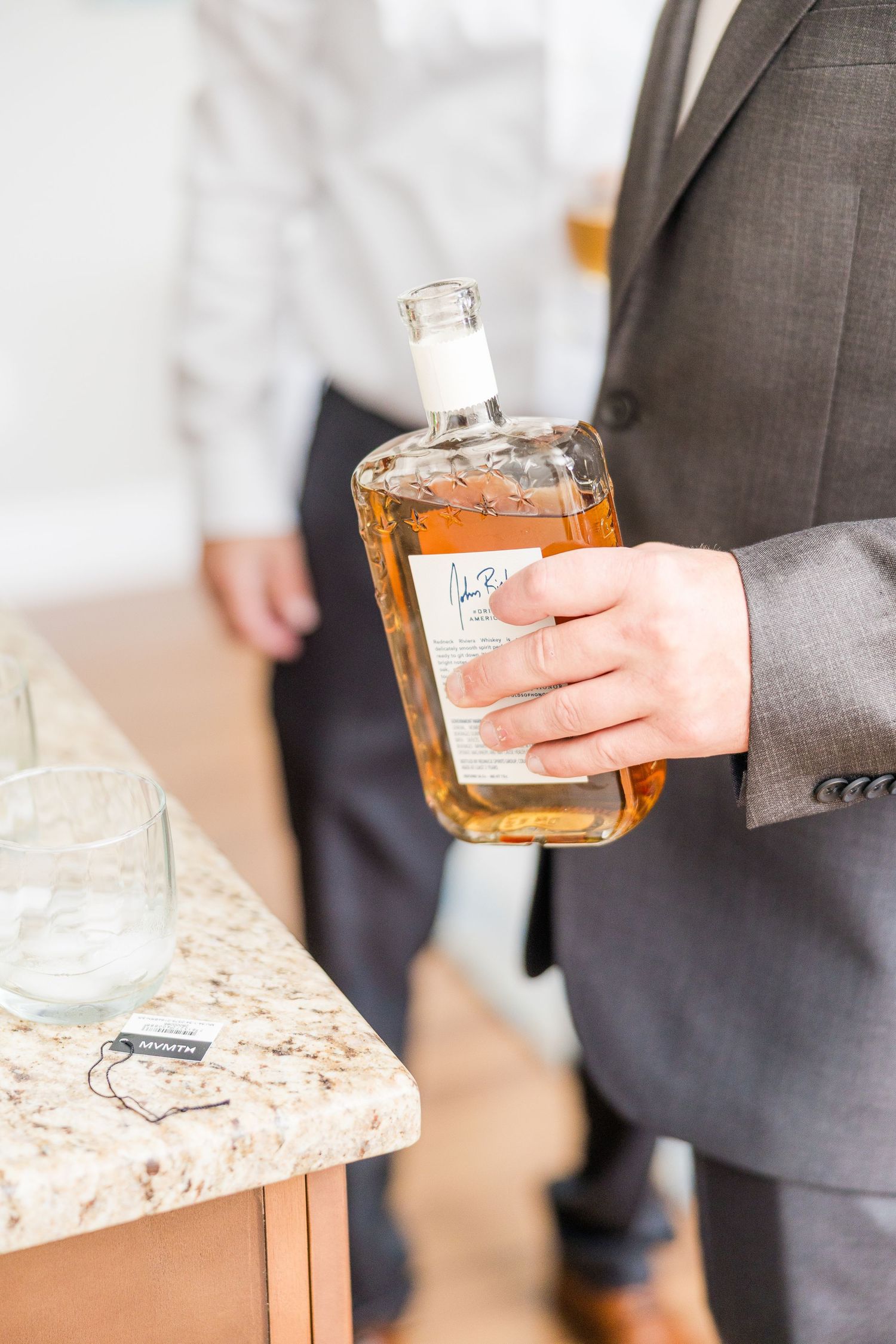 Person in grey suit holding glass of whiskey against marble countertop wedding groom bride lake Gaston photographer light airy