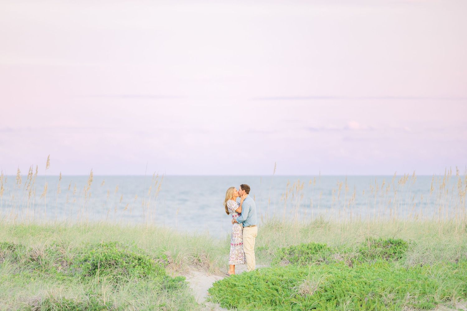 Romantic couple poses in tall grass with ocean views during sunset.