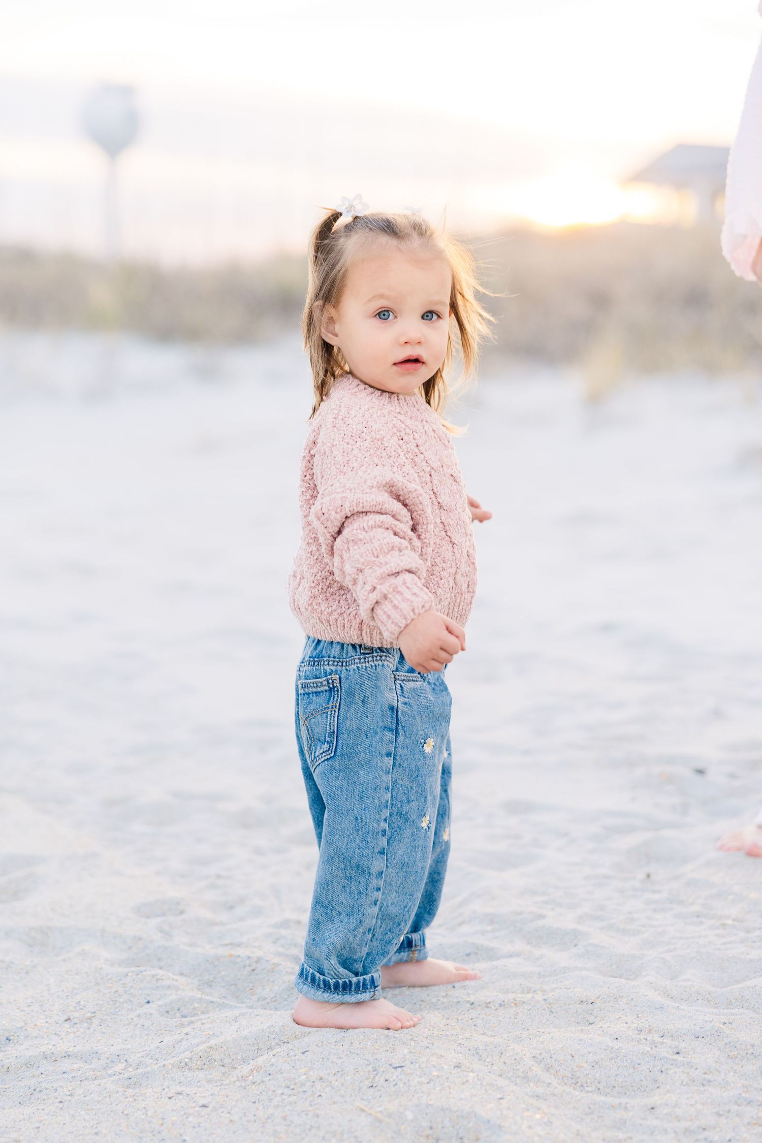 Toddler in pink sweater and denim shorts walks on white sandy beach during golden hour.