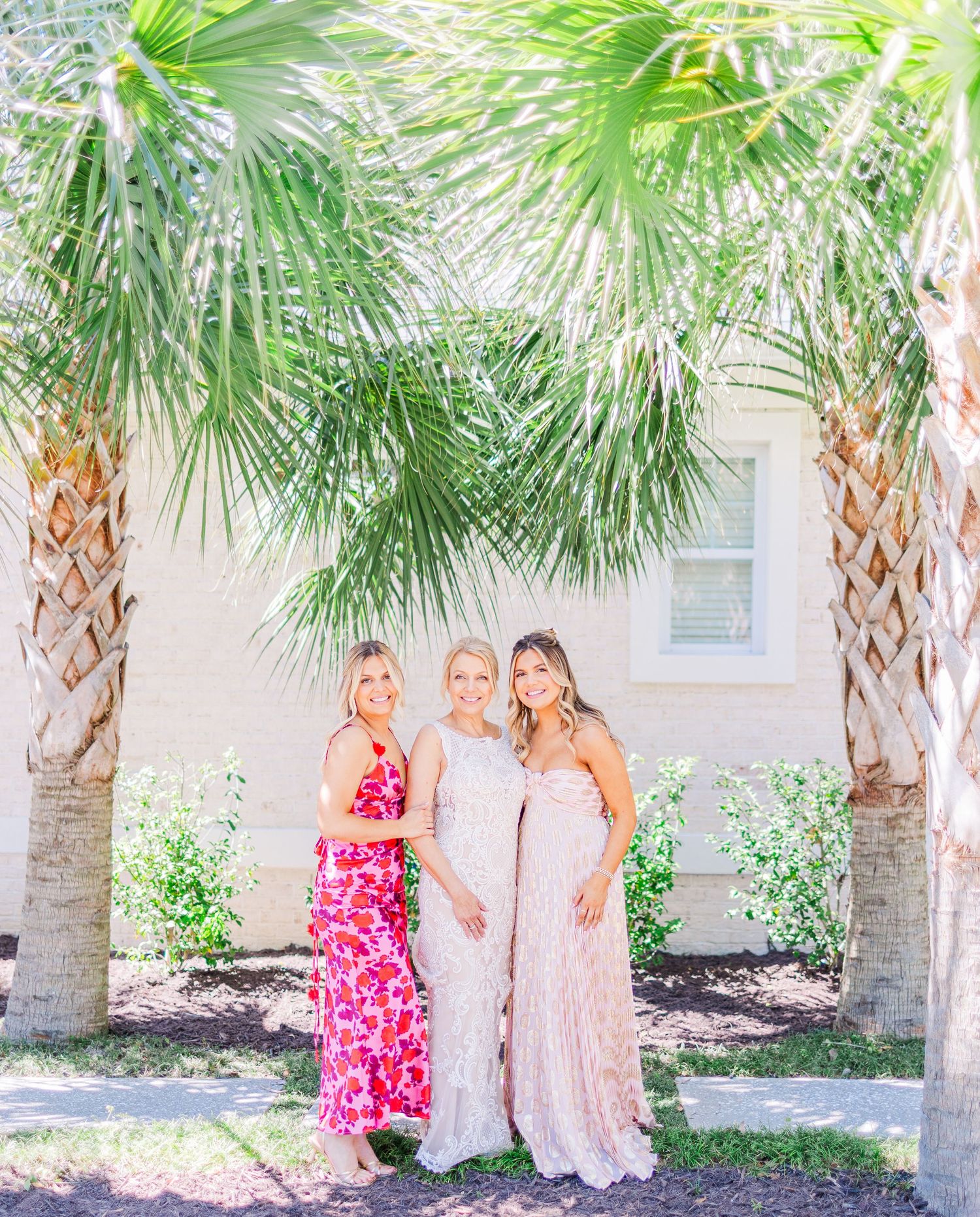 Three women in formal attire stand between palm trees for a sunny outdoor photo.