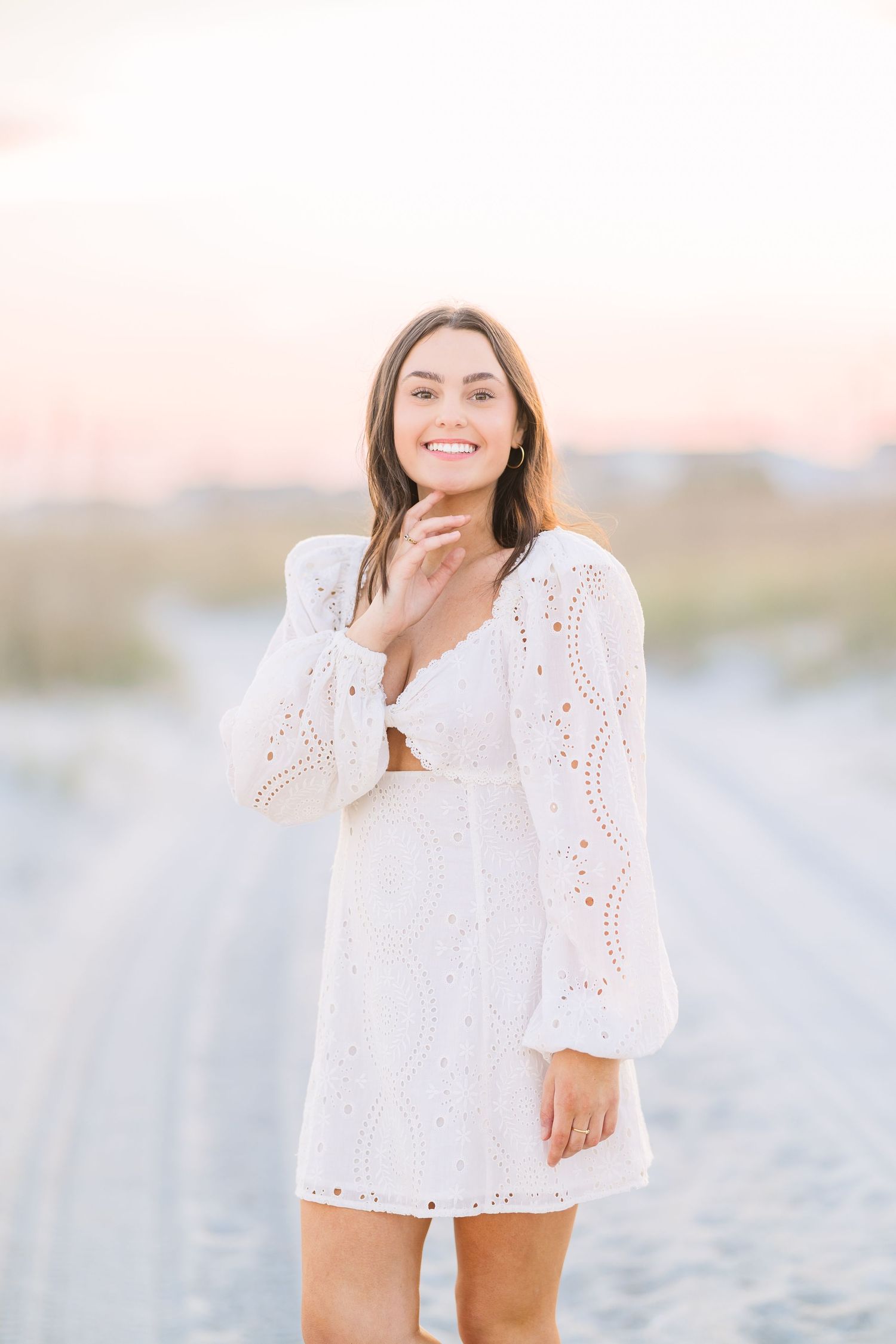 Woman in white eyelet lace dress poses on beach during sunset with soft pink sky in background.