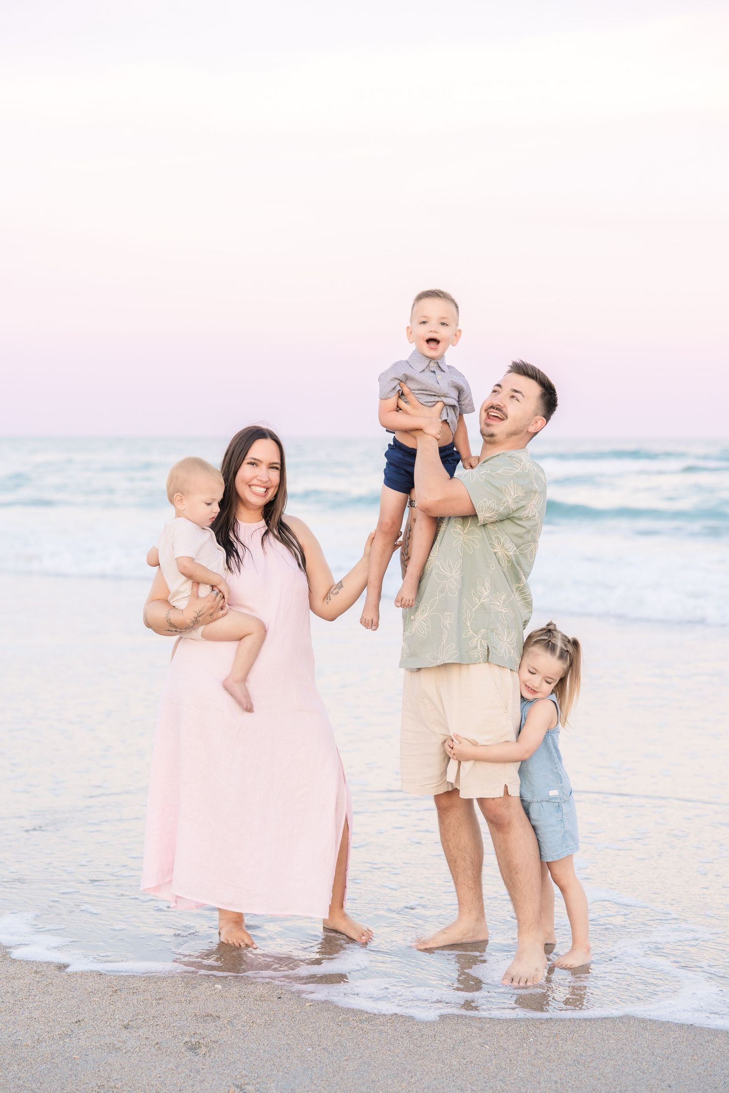 Happy family of five enjoying a sunset beach photoshoot, parents holding young children at the water's edge.