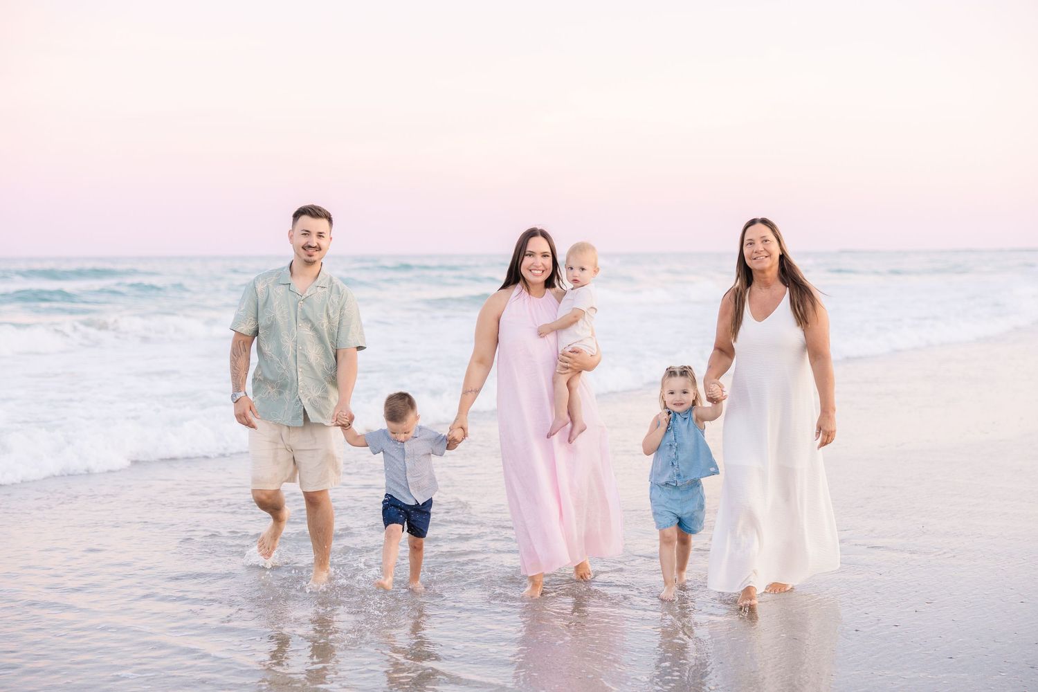 Happy family of five walking along a beach at sunset with pink sky and ocean waves.