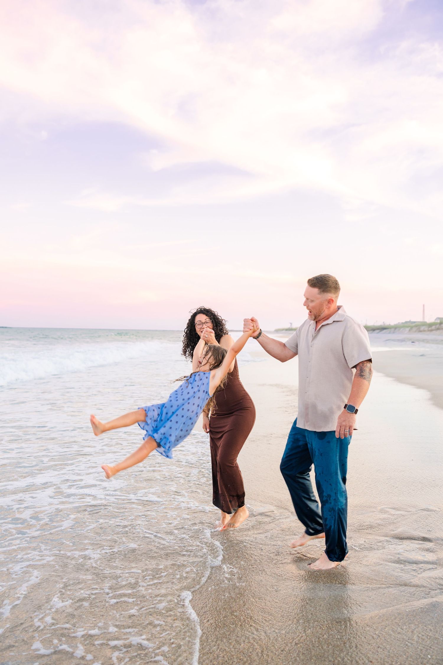 Family of three playing on the beach at sunset, parents swinging child between them near ocean waves.