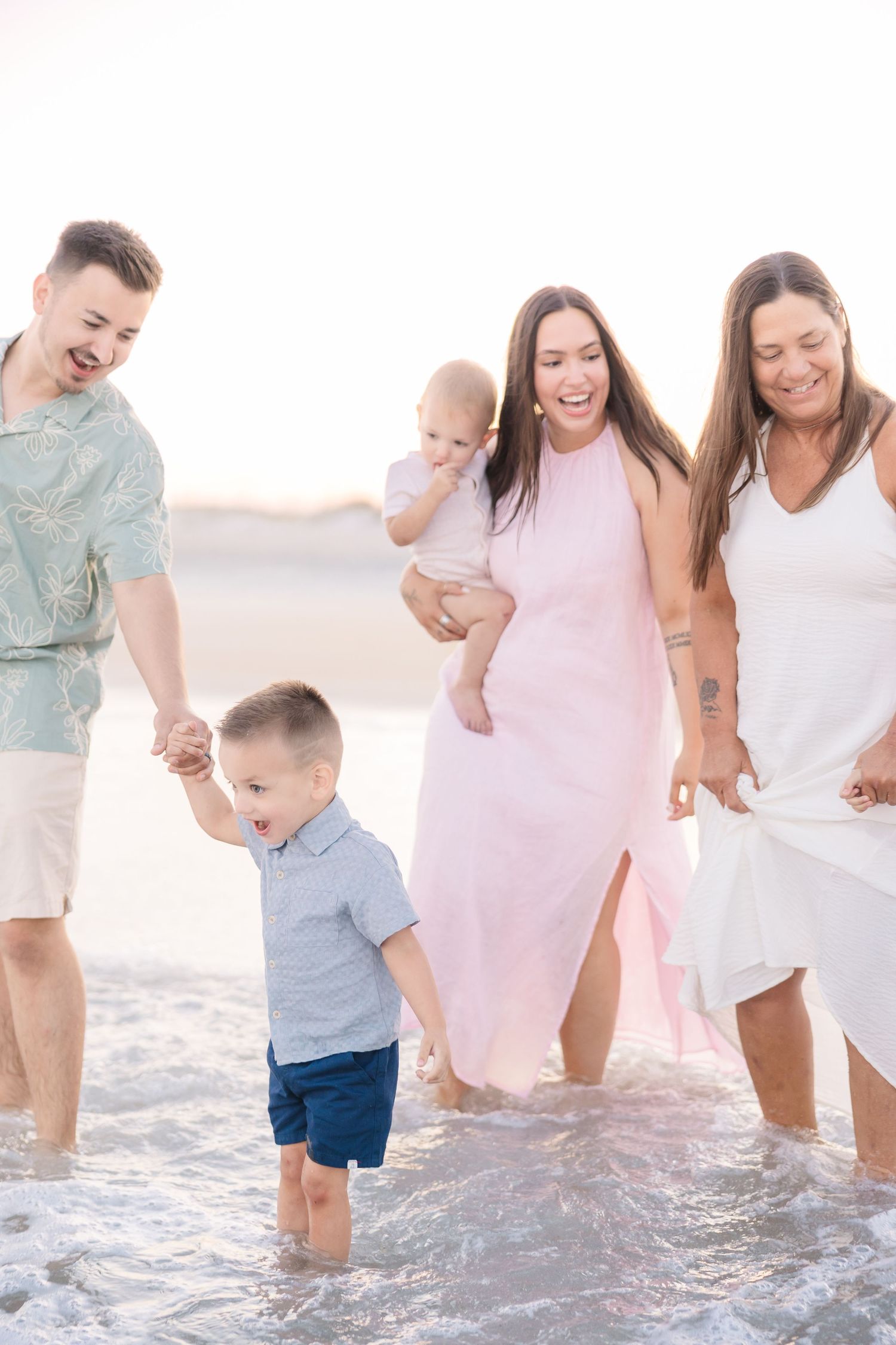 Extended family laughs and walks through shallow ocean waves together during golden hour beach session.