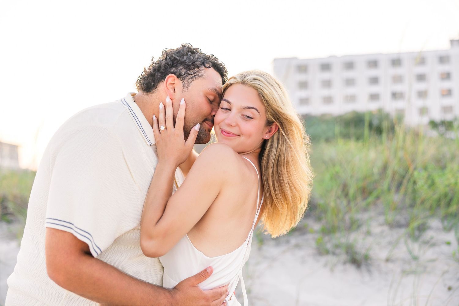 Couple sharing a tender moment on a sandy beach, man kissing woman's cheek as she smiles softly.