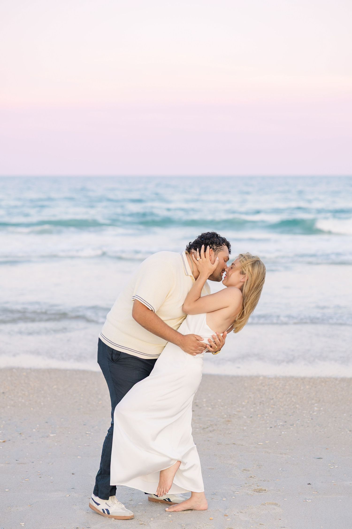 Romantic couple kissing on a beach at sunset, woman in white dress dipped back near ocean waves.