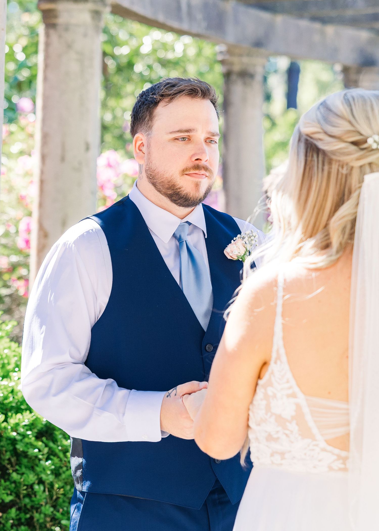 Groom in navy vest and blue tie holds bride's hands during outdoor wedding ceremony surrounded by lush greenery in Airlie Gardens in Wilming
