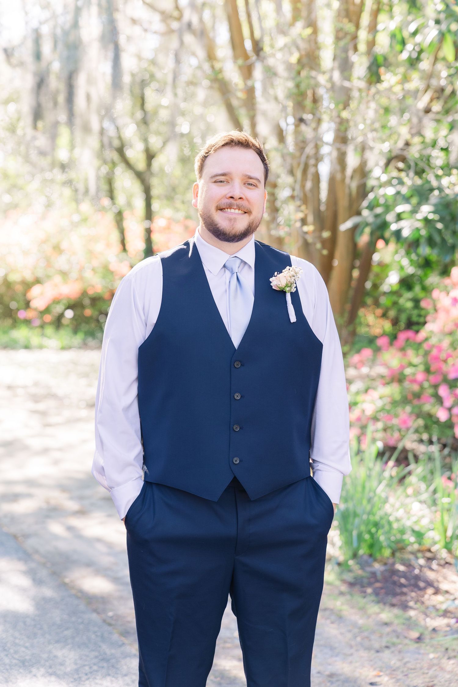 Smiling groom in navy vest and light blue tie with boutonniere poses outdoors among colorful spring garden in Airlie Gardens in Wilmington,