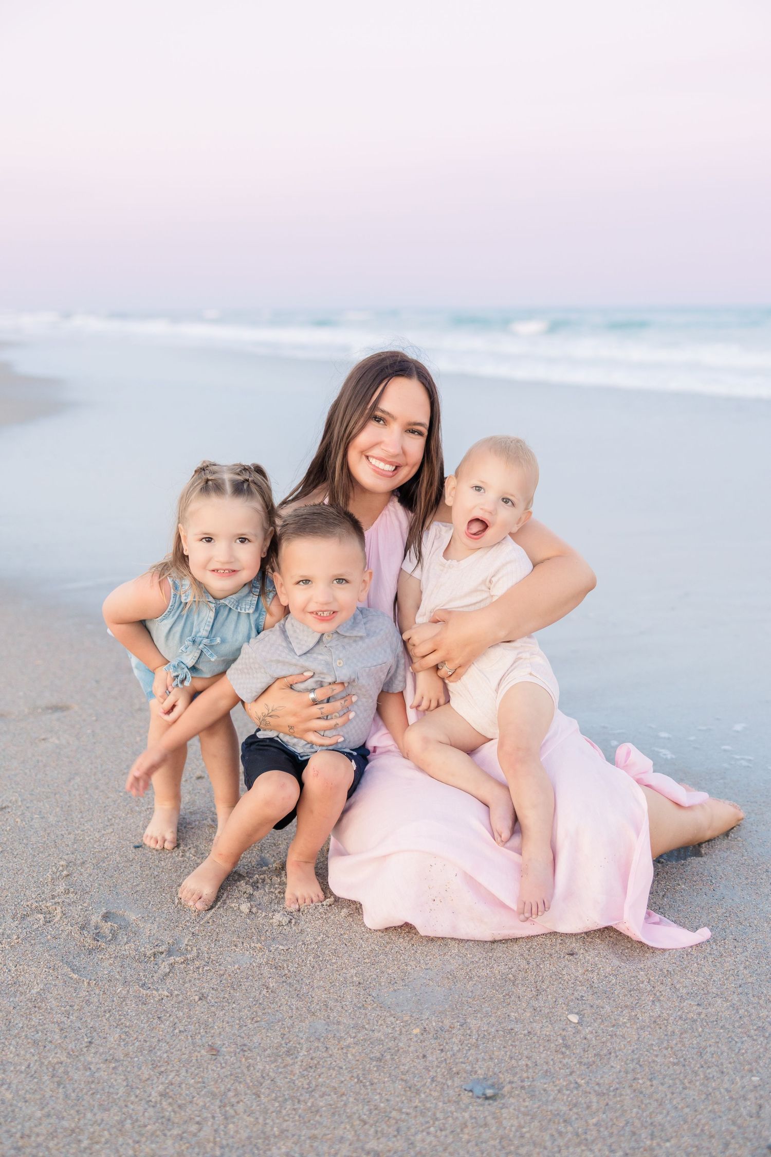 A smiling mother sits on a sandy beach holding her baby while her two young children cuddle close beside her.