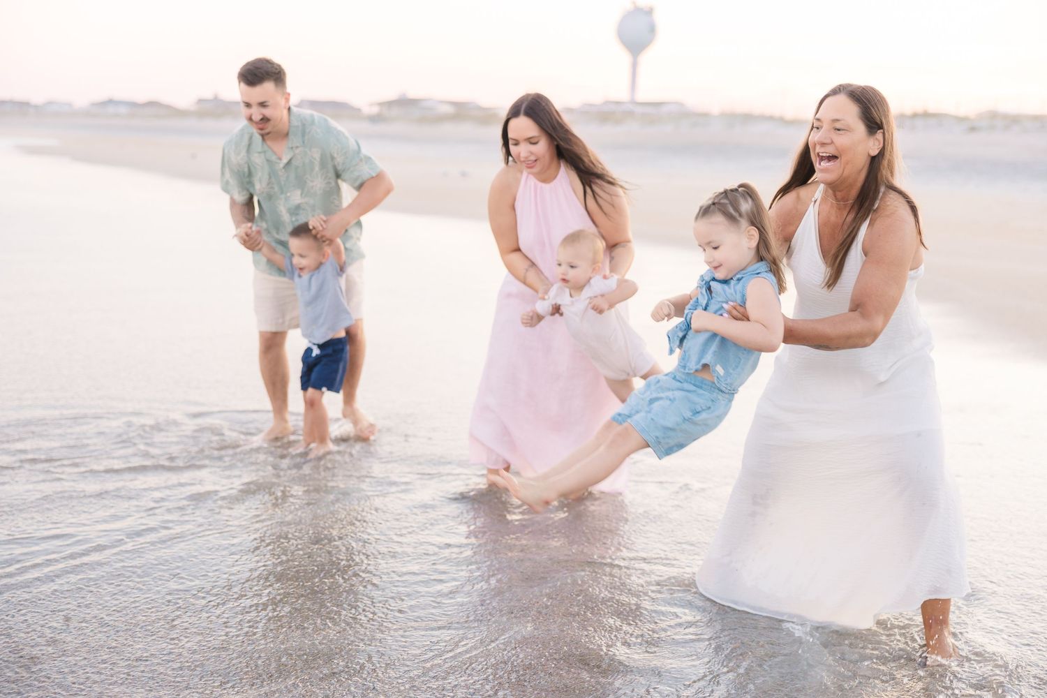 Two families laugh and play with toddlers in shallow ocean waves on a sandy beach at golden hour.