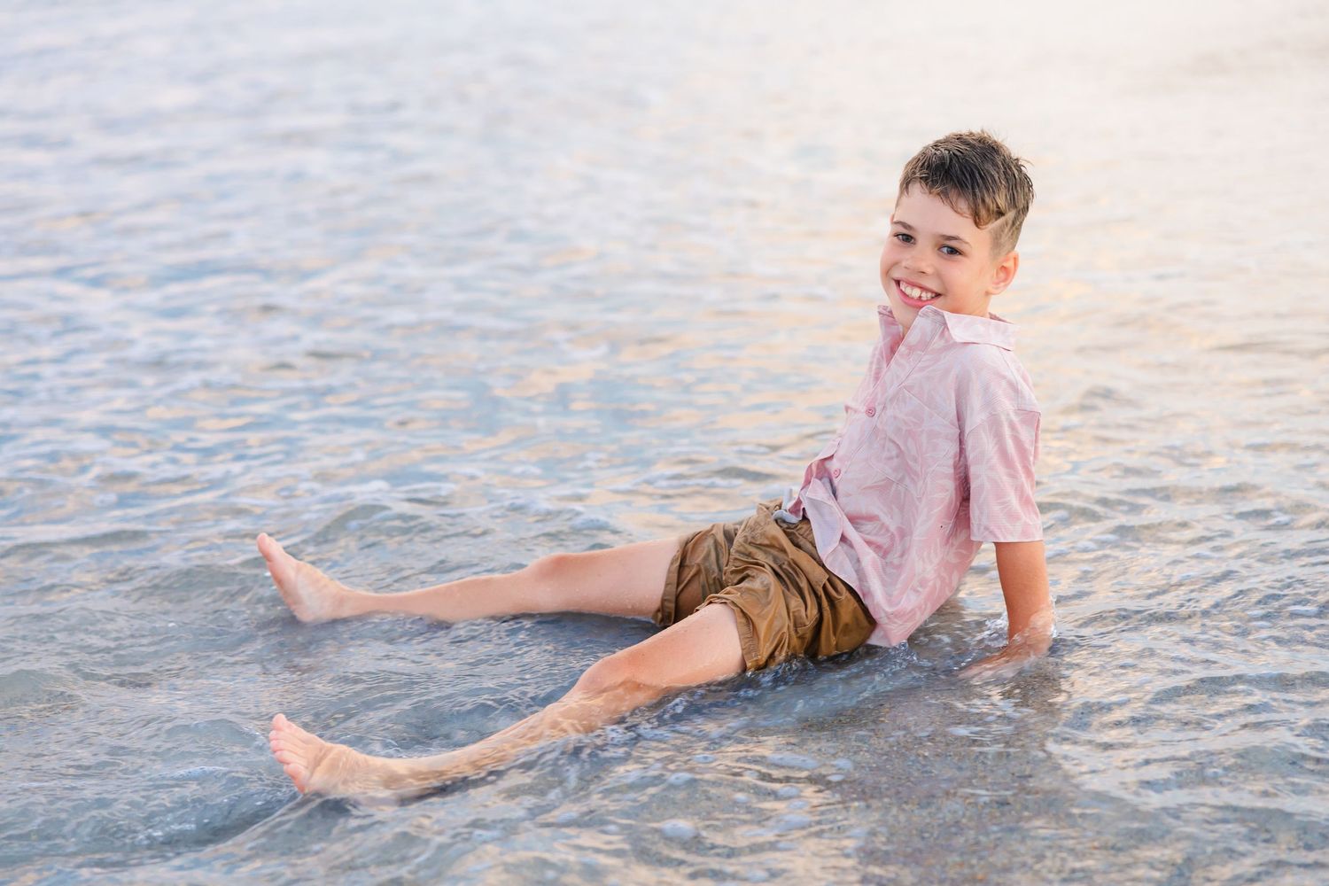 A smiling boy in a pink shirt and khaki shorts sits in shallow ocean water at the beach.