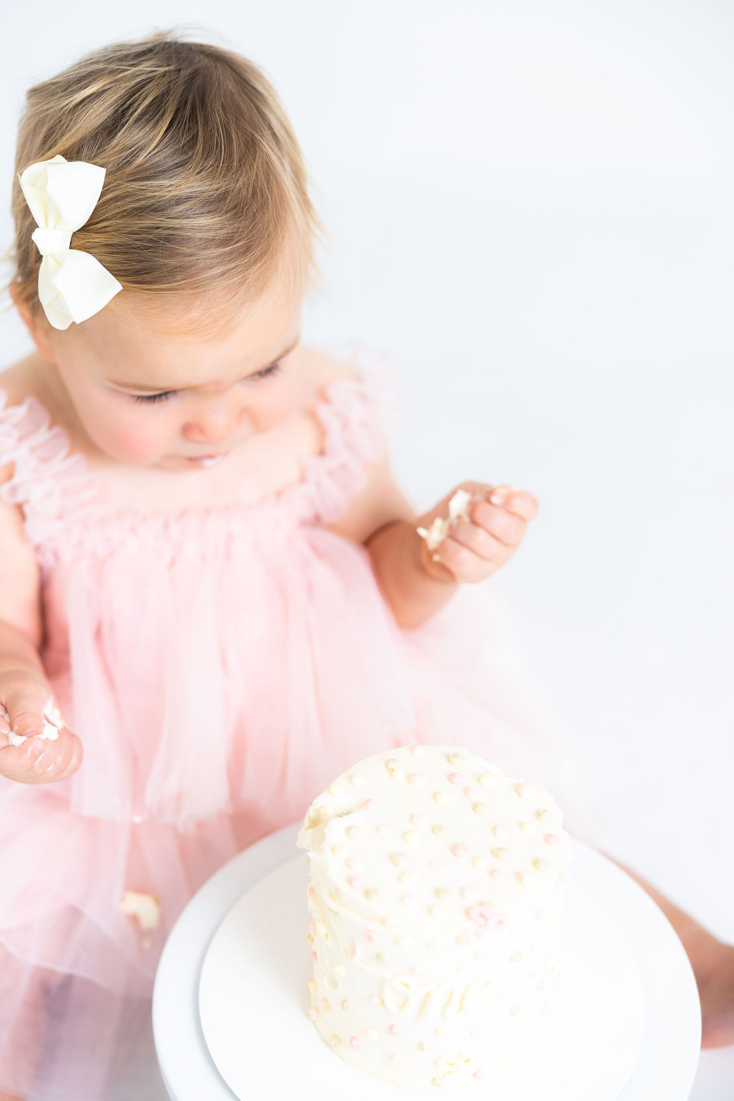 Baby in pink outfit leans over to touch white frosted cake on stand.