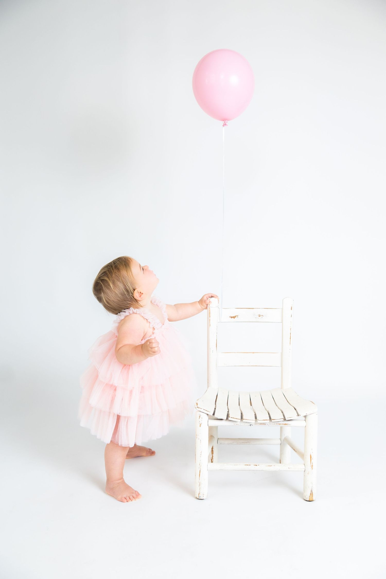 Baby in pink dress reaching towards white chair and pink balloon on white backdrop.