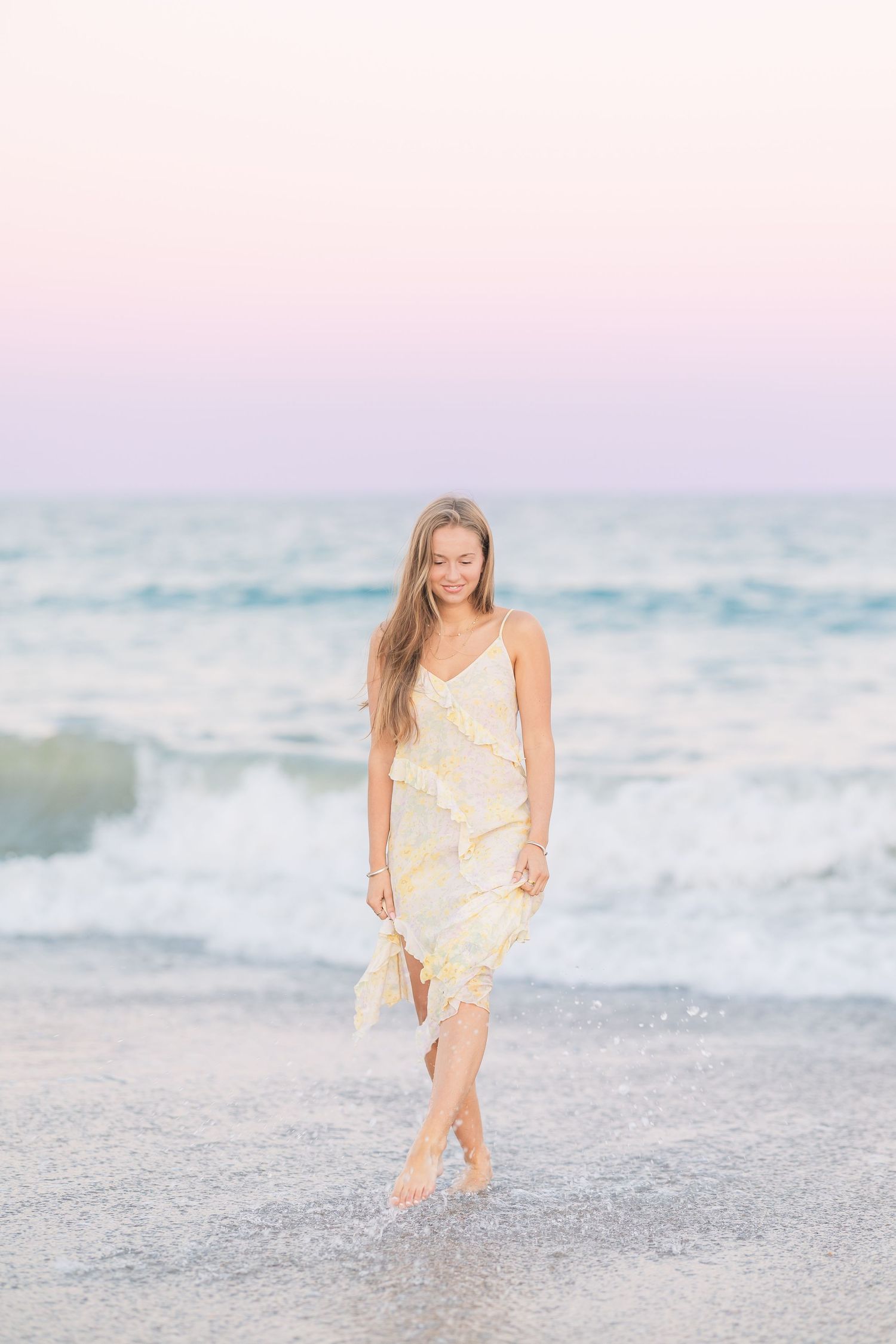 A person in a yellow dress plays in the ocean waves at sunset with dreamy pastel colors.