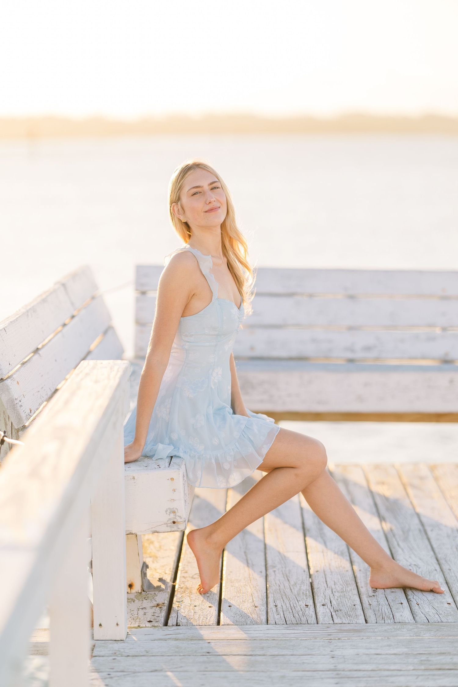 Relaxed summer pose on weathered white wooden bench in light blue dress at sunset.