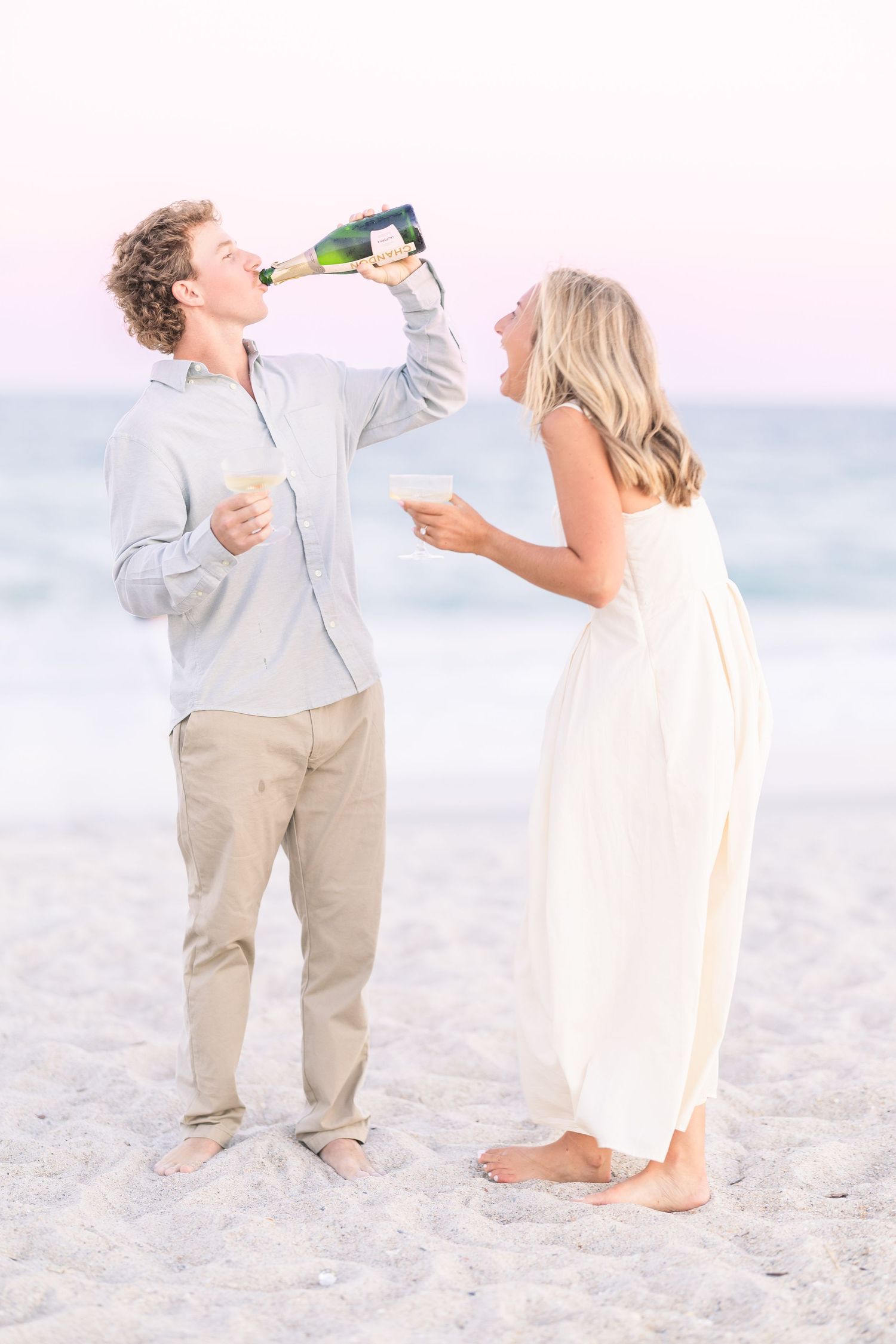 A couple celebrates with champagne on a bright sandy beach while wearing white and beige attire.