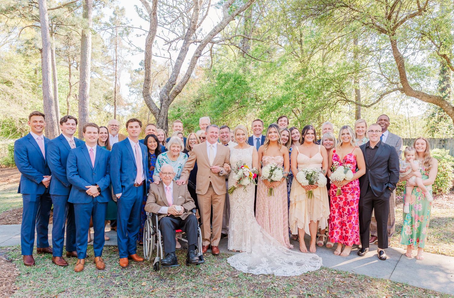 Wedding party group photo with bridesmaids in blush dresses and groomsmen in navy suits outdoors.