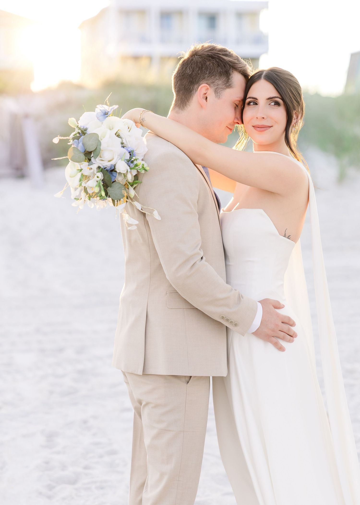A bride and groom embrace on a sandy beach at sunset, she holds a blue and white bouquet while wearing a strapless gown.