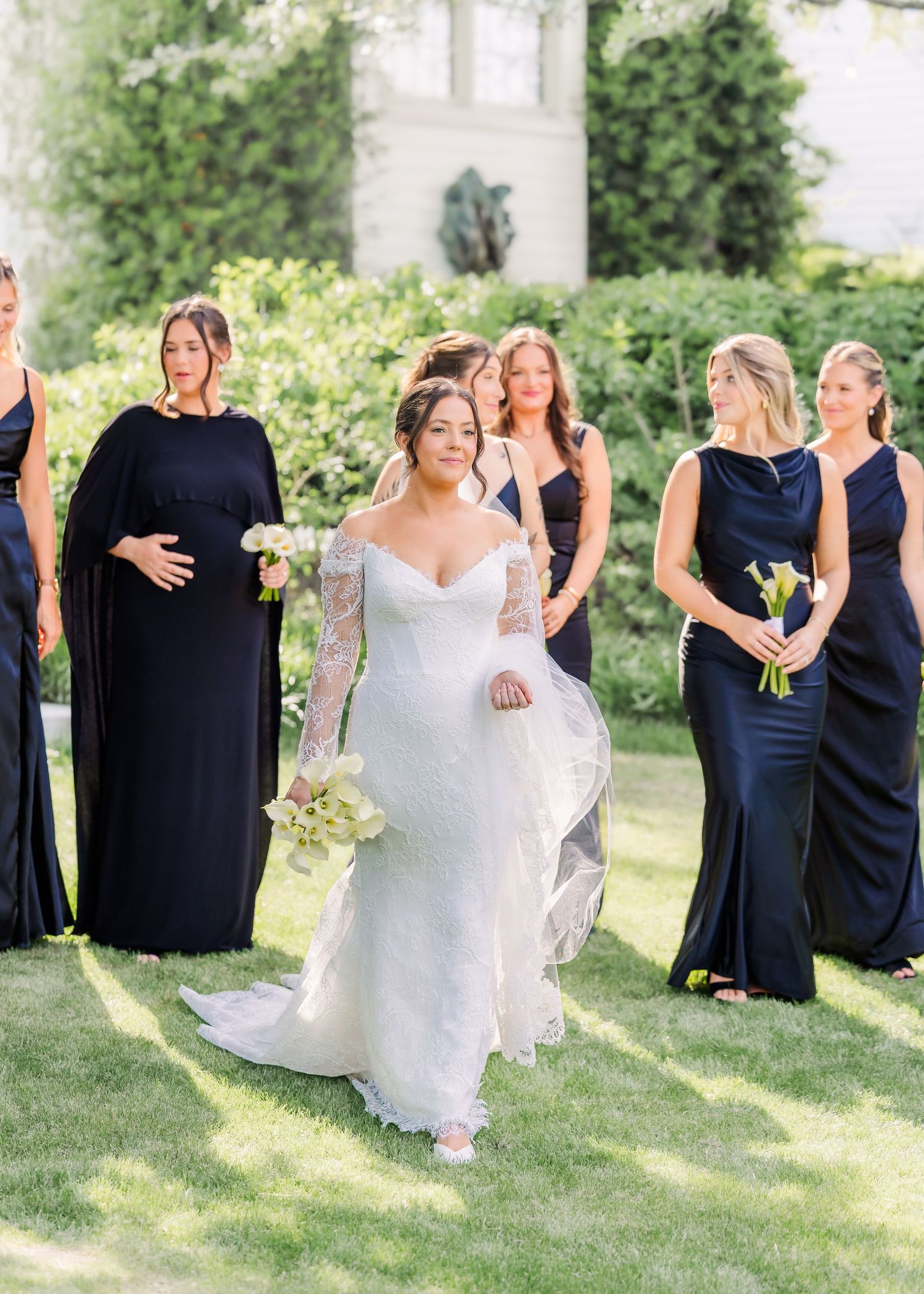 Bride in white lace gown walks with bridesmaids in navy dresses on green lawn at outdoor wedding.