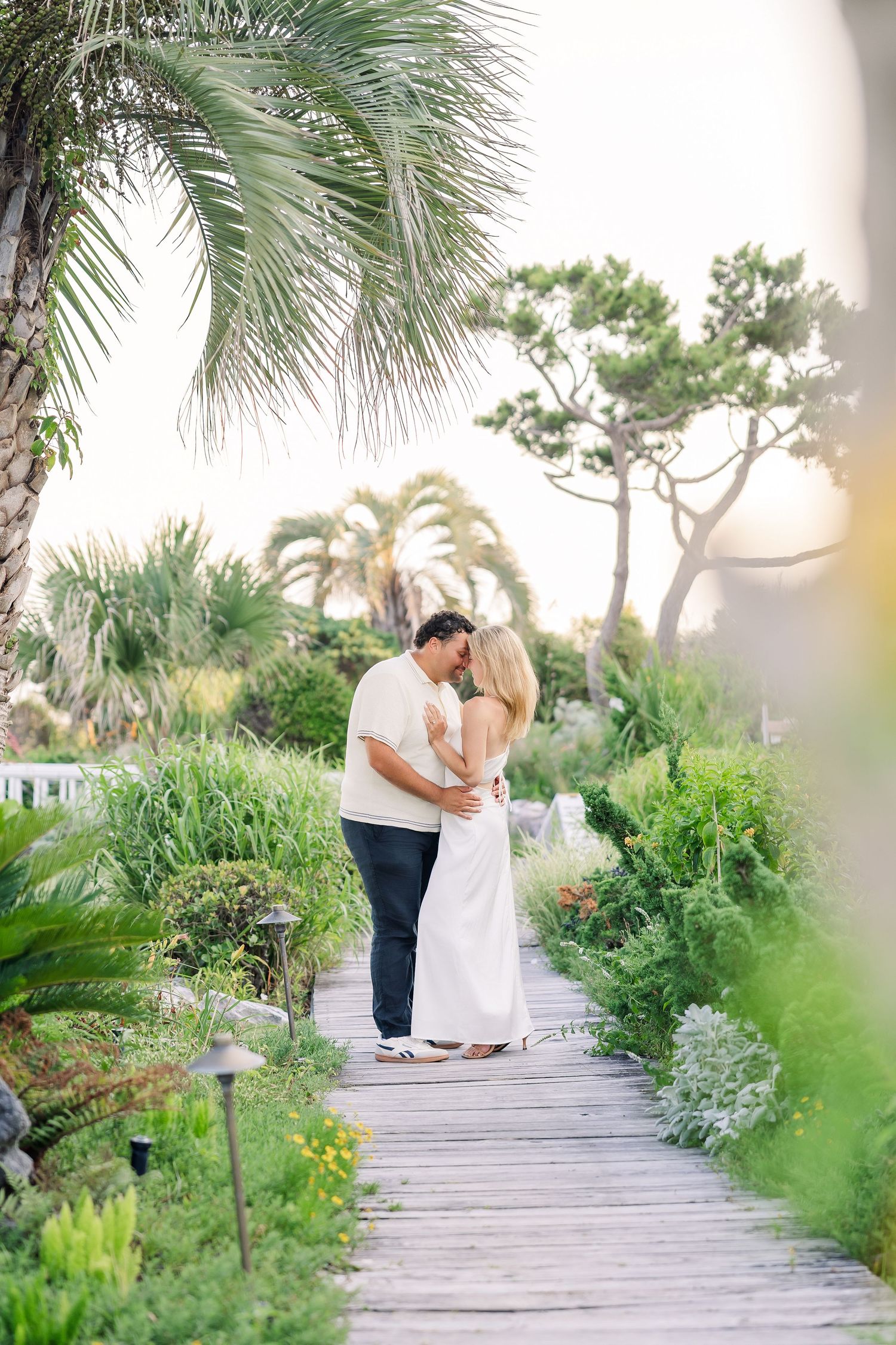 Couple embracing on a wooden boardwalk surrounded by tropical palm trees and lush garden greenery.