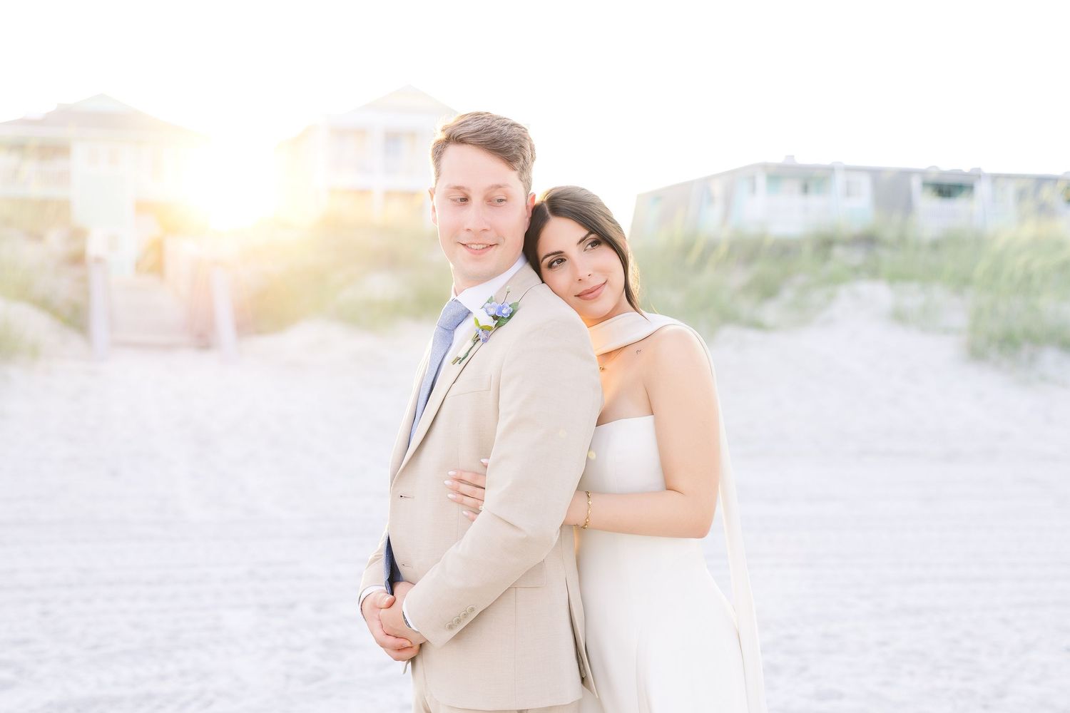 Bride and groom embracing on a sandy beach at golden hour, groom in tan suit, bride in white dress in Carolina Beach near Wilmington, NC.